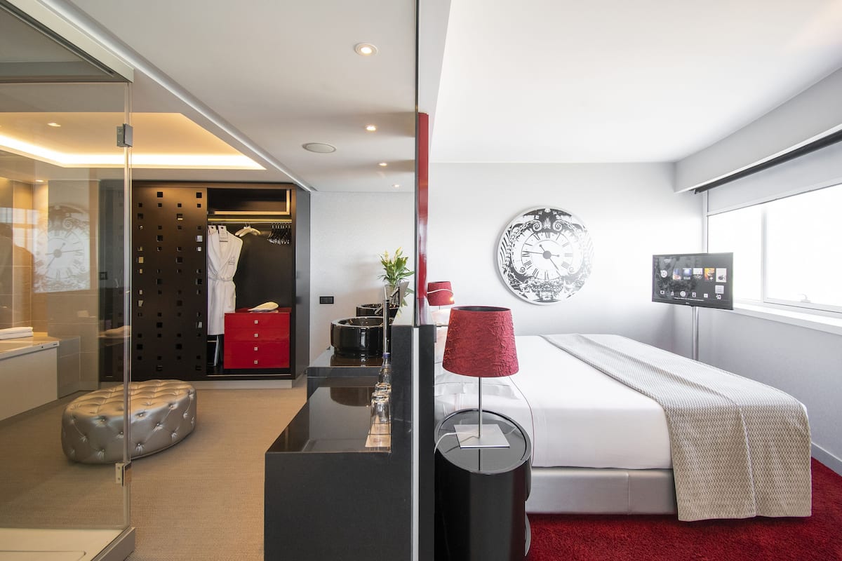 A modern bedroom with a bed, a bedside table, a red lamp, a wall clock, and a window.
