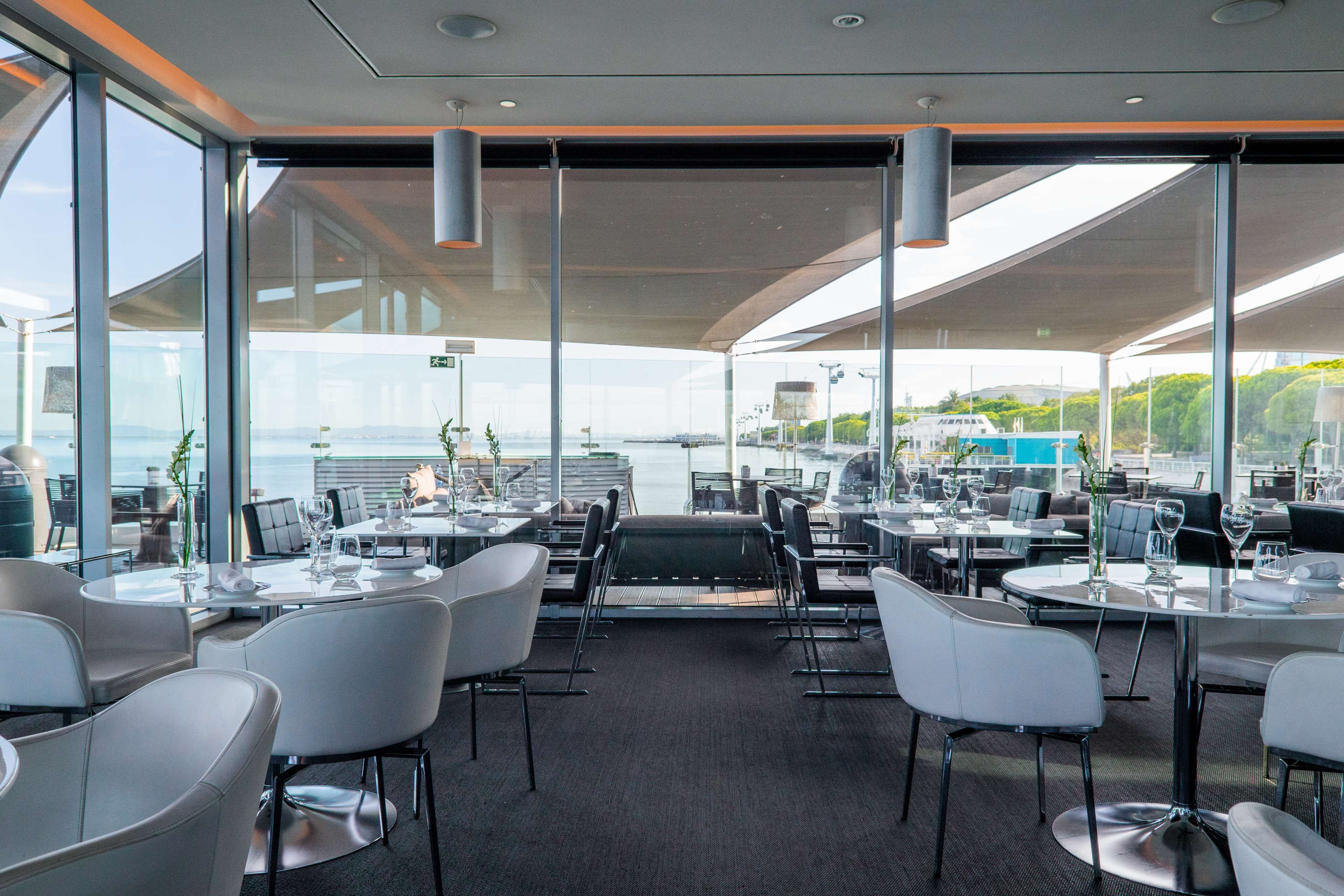 Un restaurante moderno con vista al mar, con mesas redondas, sillas blancas y un panel de vidrio.