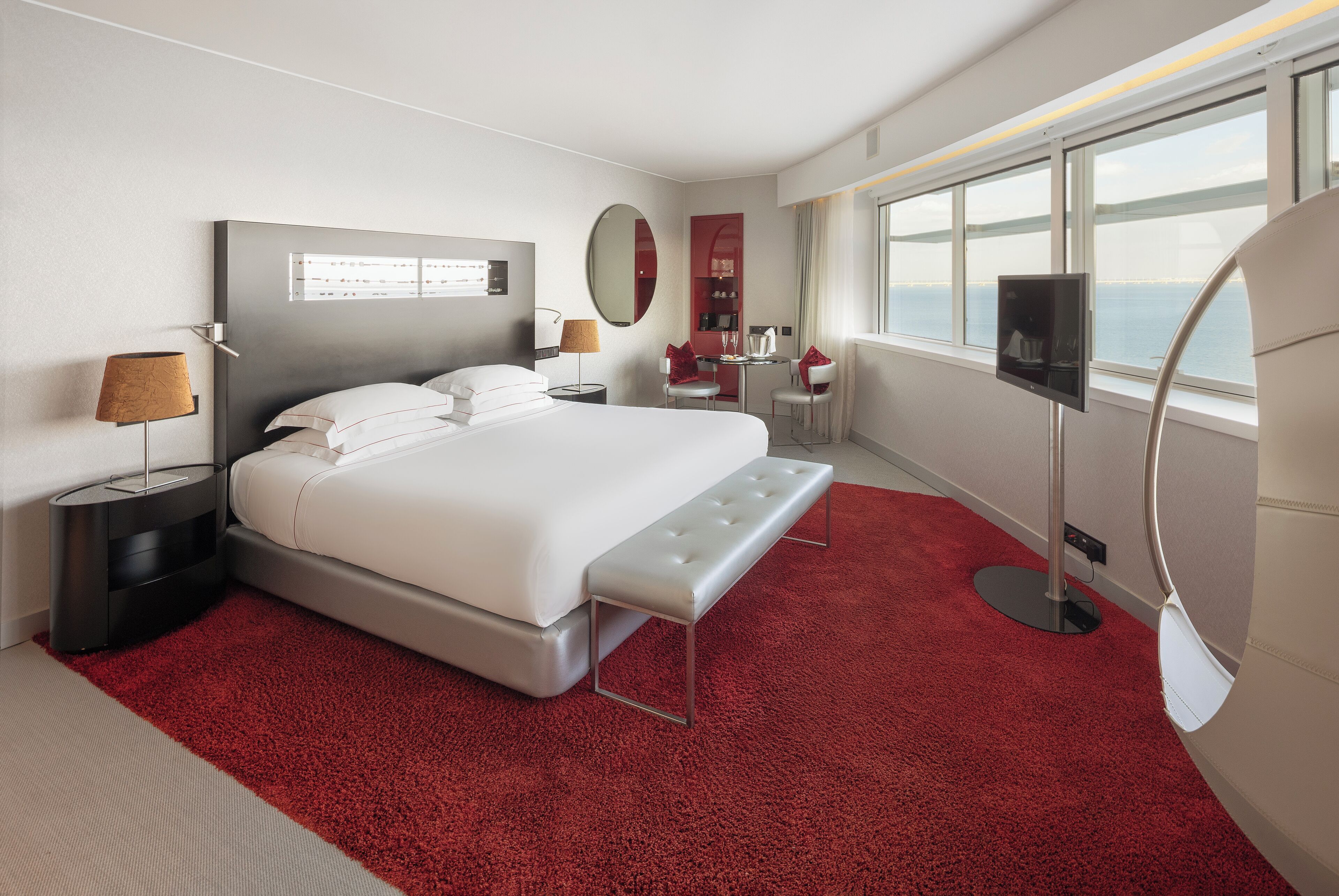 A modern hotel room with a large bed, a desk, a chair, a TV, and a view of the sea.