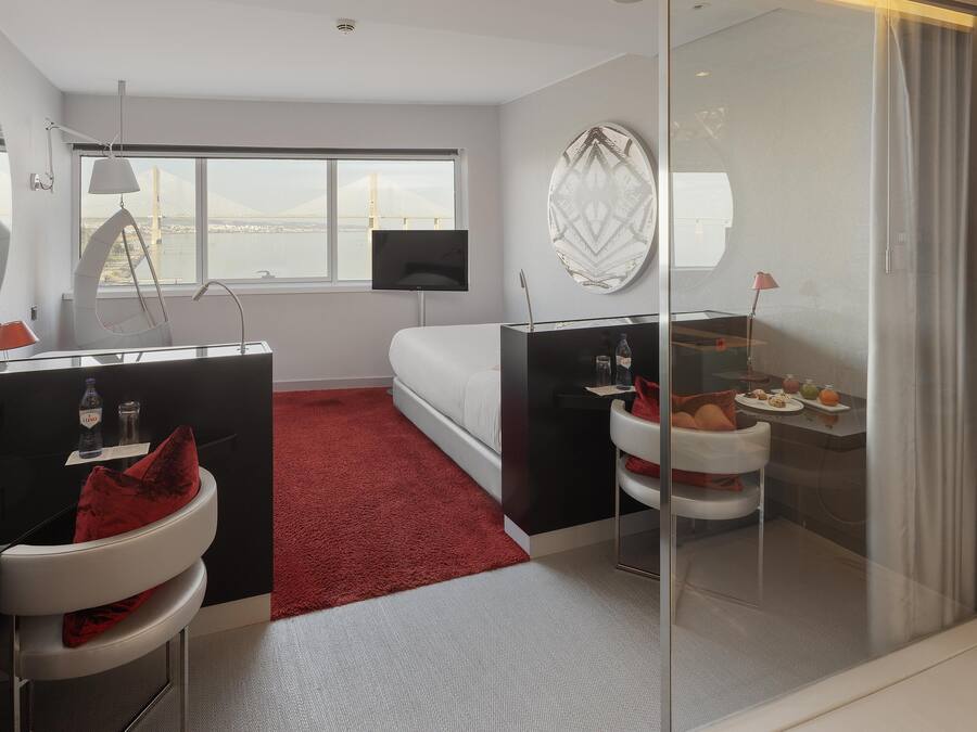A modern hotel room with a bed, a desk with a computer, a glass shower enclosure, and a view of a bridge.