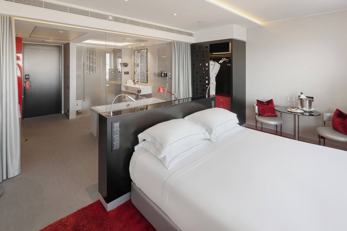 A modern hotel room with a large bed, a vanity with a mirror, a small table with a red pillow, and a red rug.