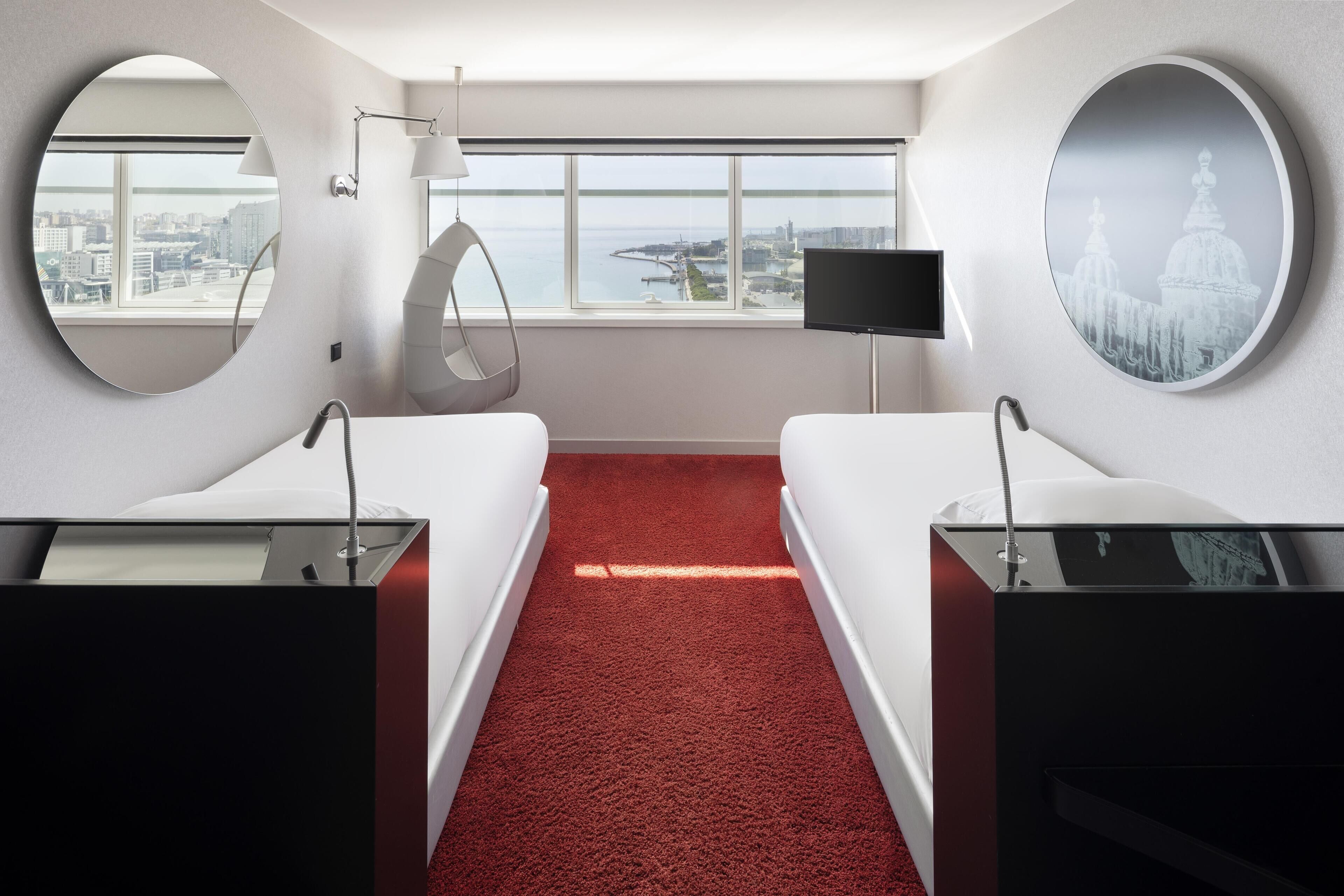 A hotel room with a red carpet, two beds, a TV, and a large window with a city view.