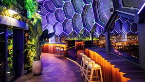 Un espace bar moderne avec des panneaux hexagonaux au plafond, un comptoir en bois et une vue sur le paysage urbain.