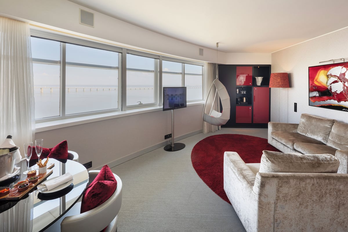 A modern hotel room with a large window, a TV, a red carpet, and a seating area with a sofa and chairs.