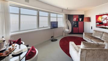 Una habitación de hotel moderna con un gran ventanal, televisión, alfombra roja y una zona de estar con sofá y sillones.