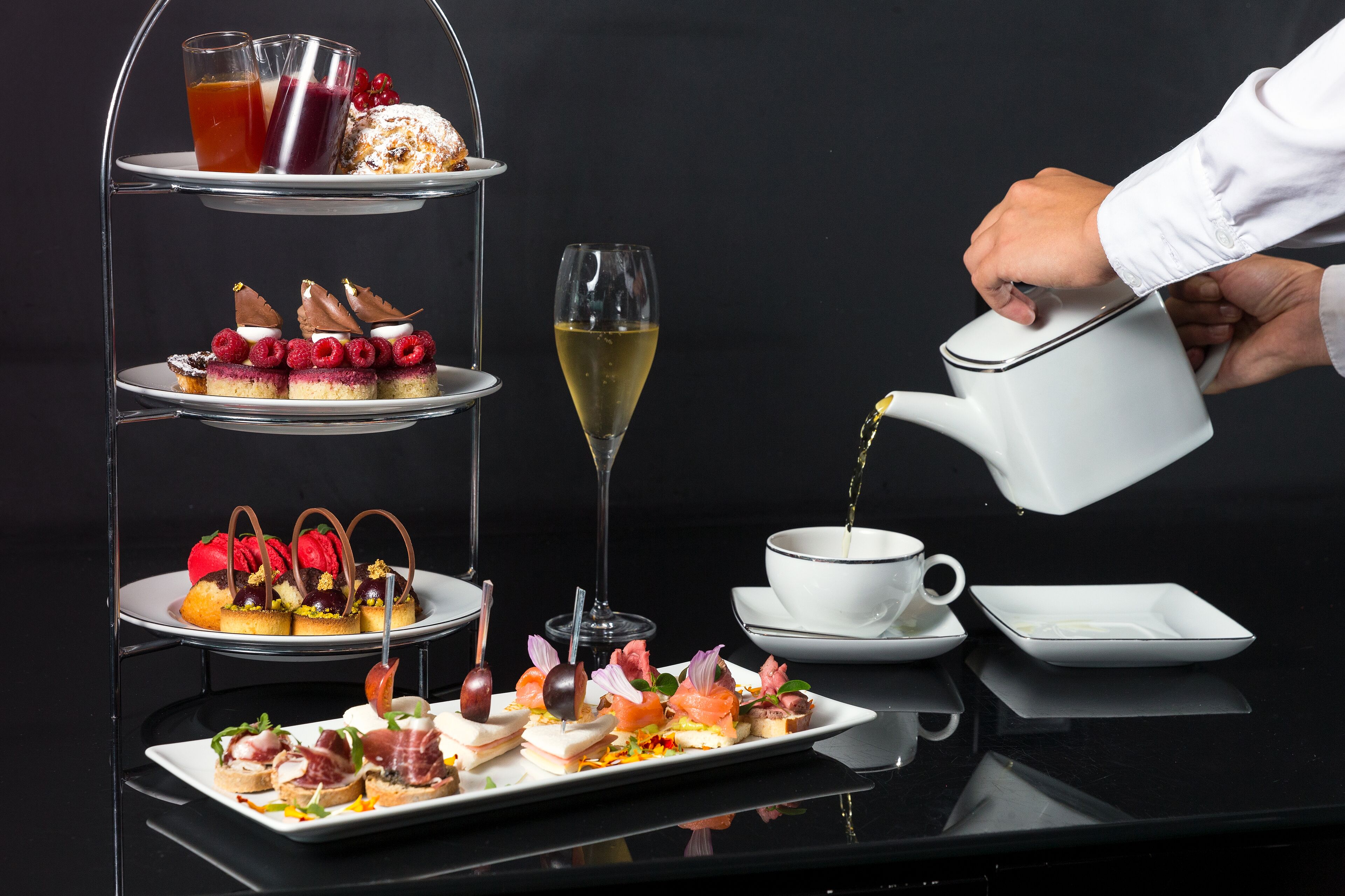 A three-tiered dessert stand with various pastries and a glass of champagne. A hand is pouring a drink into a cup.