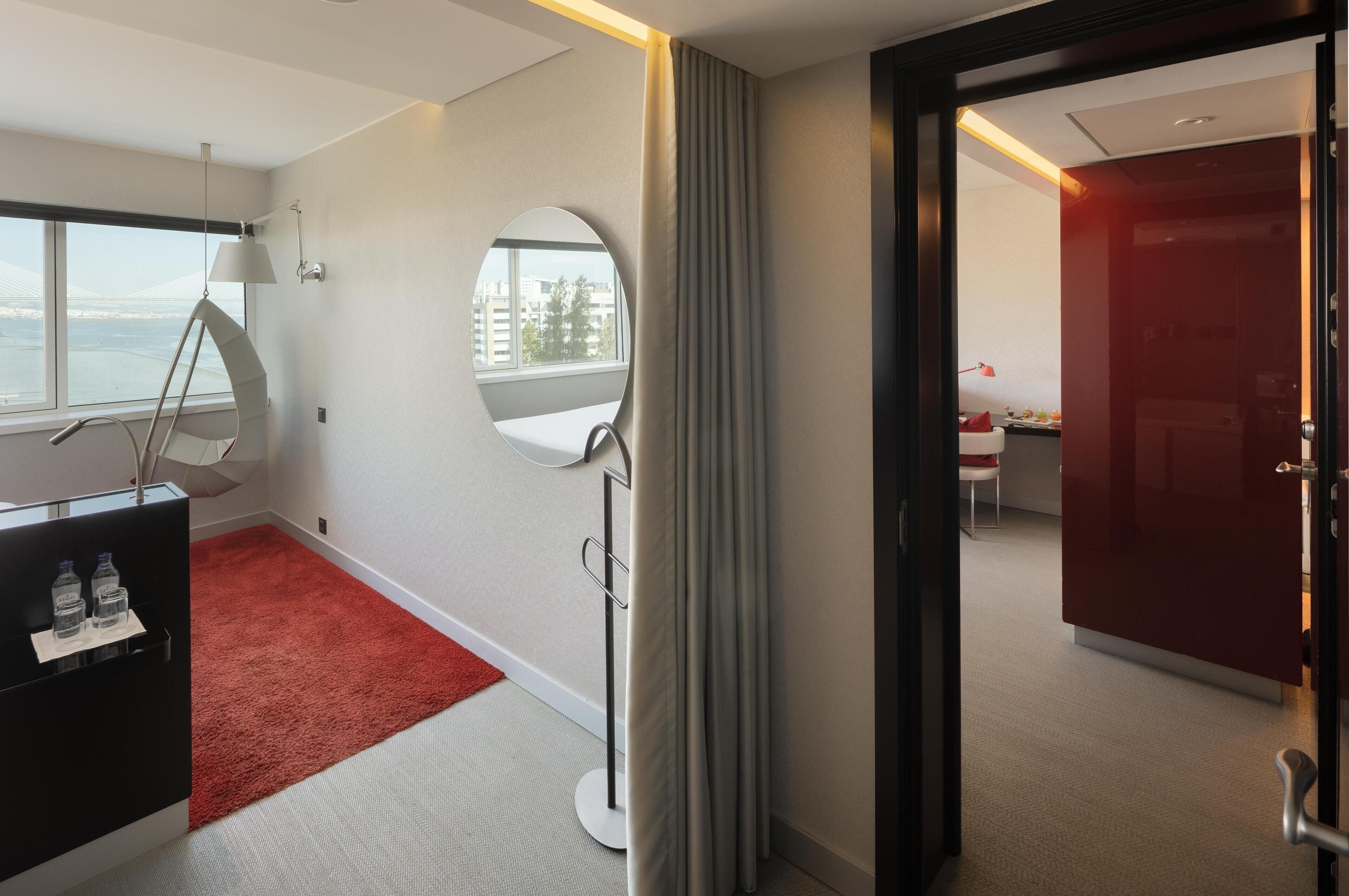 Habitación de hotel con un ventanal grande, un escritorio con espejo, alfombra roja y puerta roja.