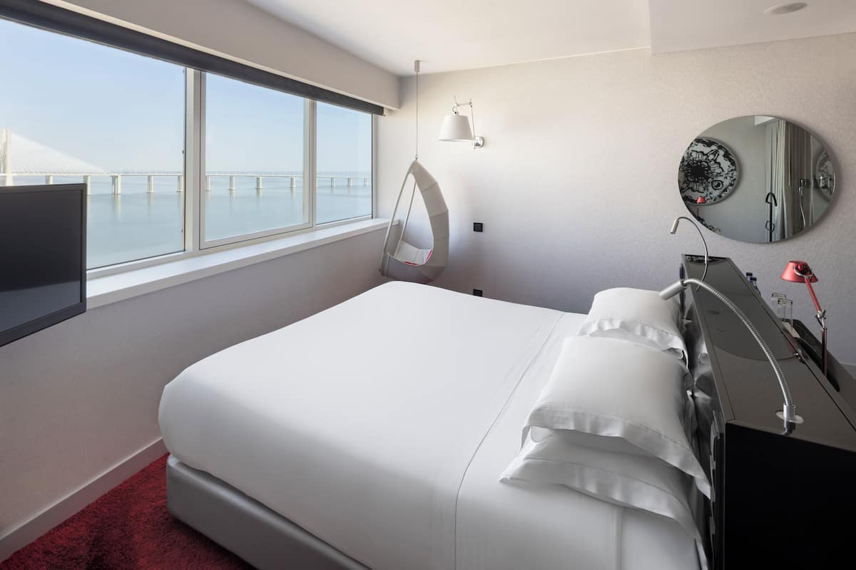 A modern hotel room with a large bed, a TV, a hanging chair, a round mirror, and a view of a bridge.