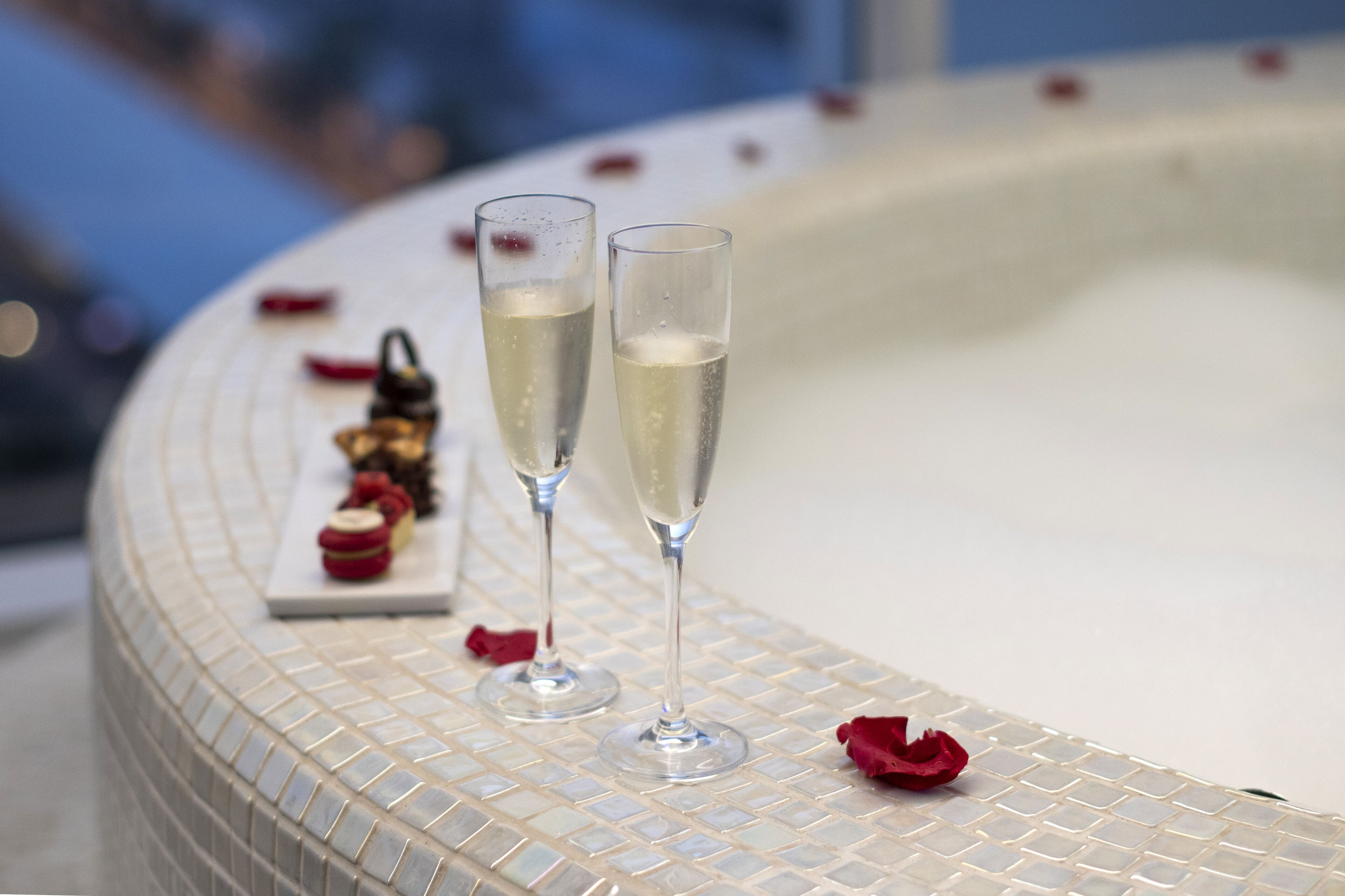 Two champagne flutes on a tiled surface with a small tray of desserts and rose petals.
