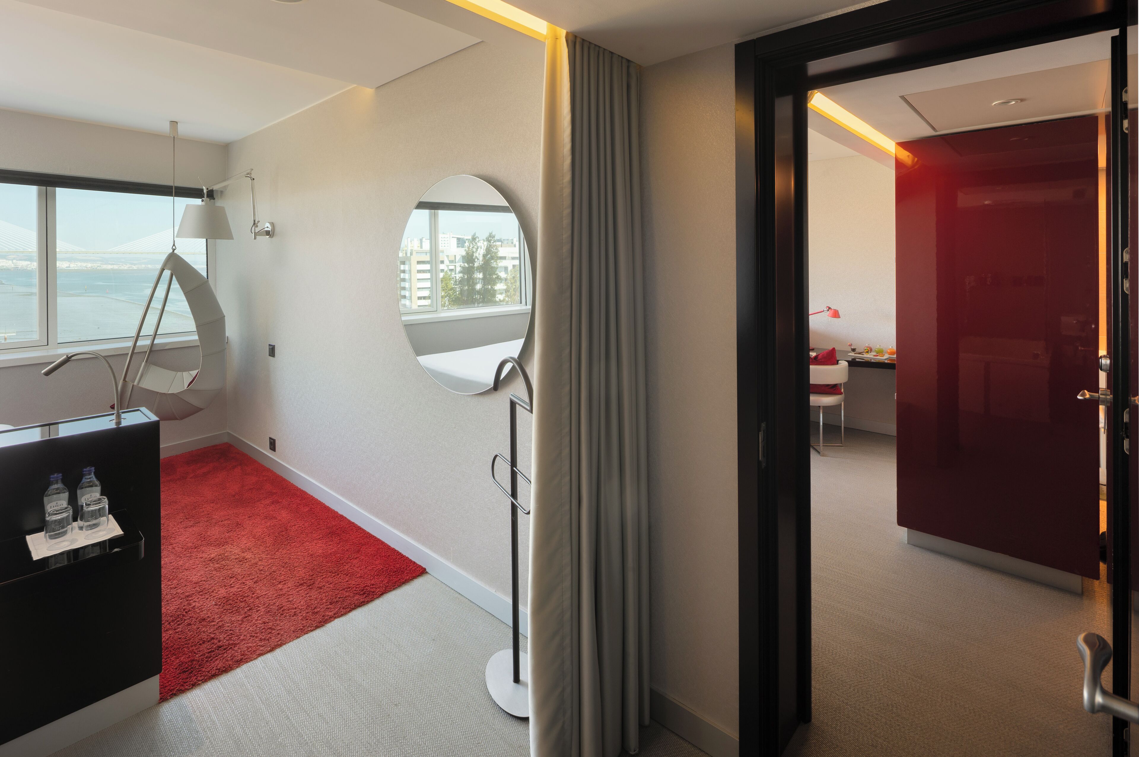 A hotel room with a red carpet, a desk with a mirror, a hanging chair, and a view of the city through large windows.