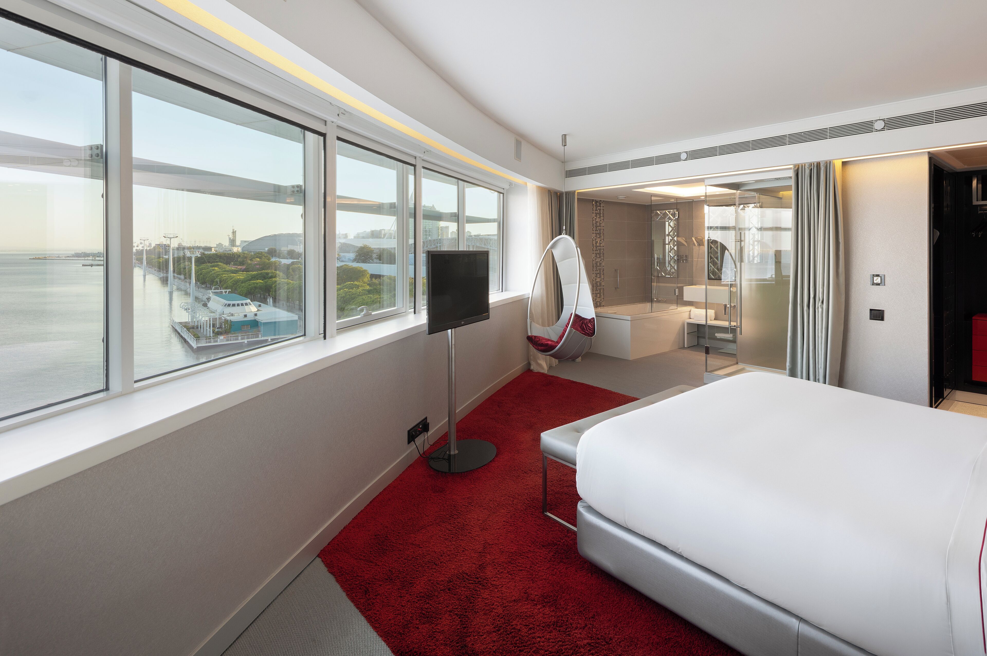 A hotel room with a large window, a bed, a TV, a red carpet, and a bathroom with a glass shower.