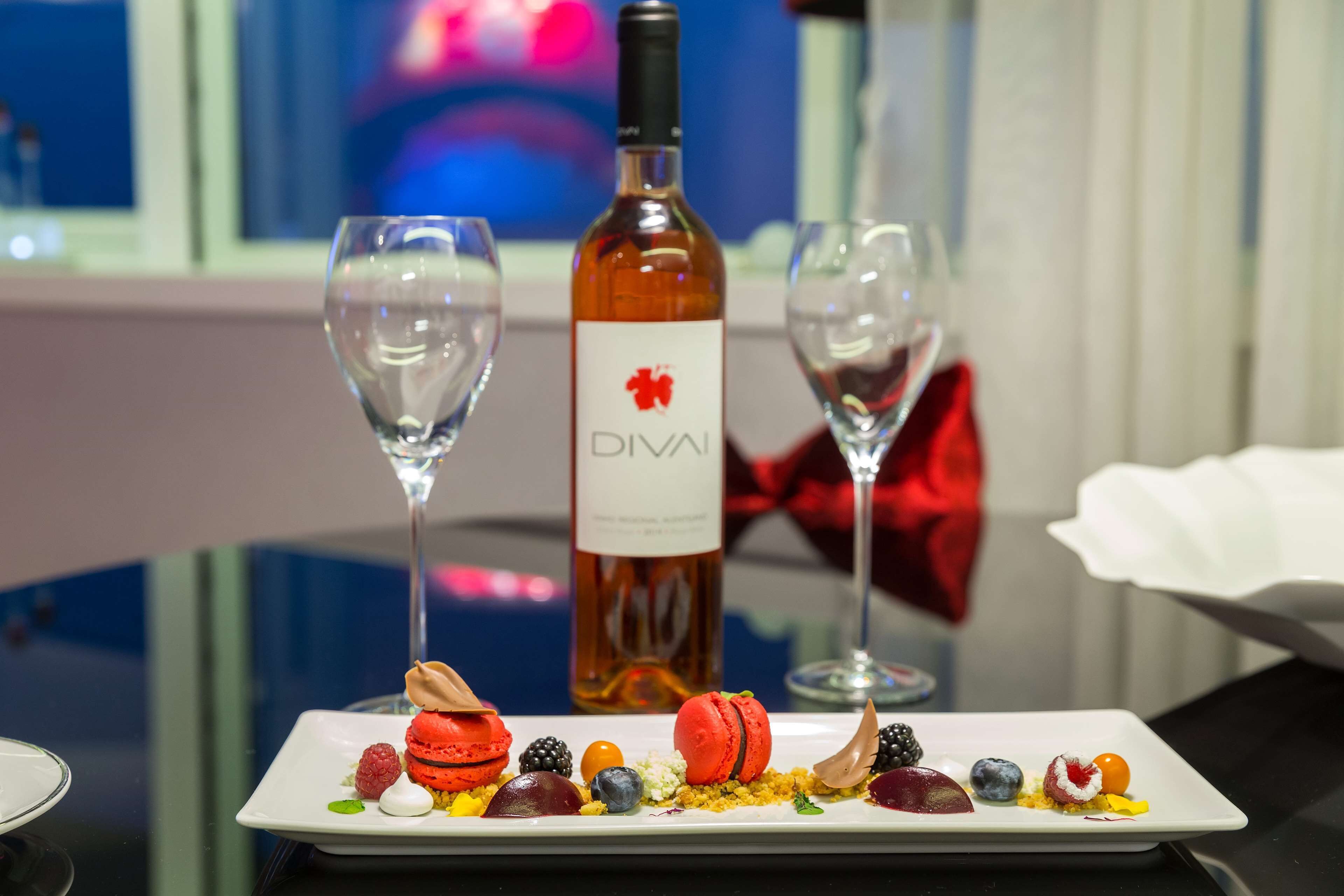 A dessert plate with macarons and berries, a bottle of Divali wine, and two empty wine glasses on a table.
