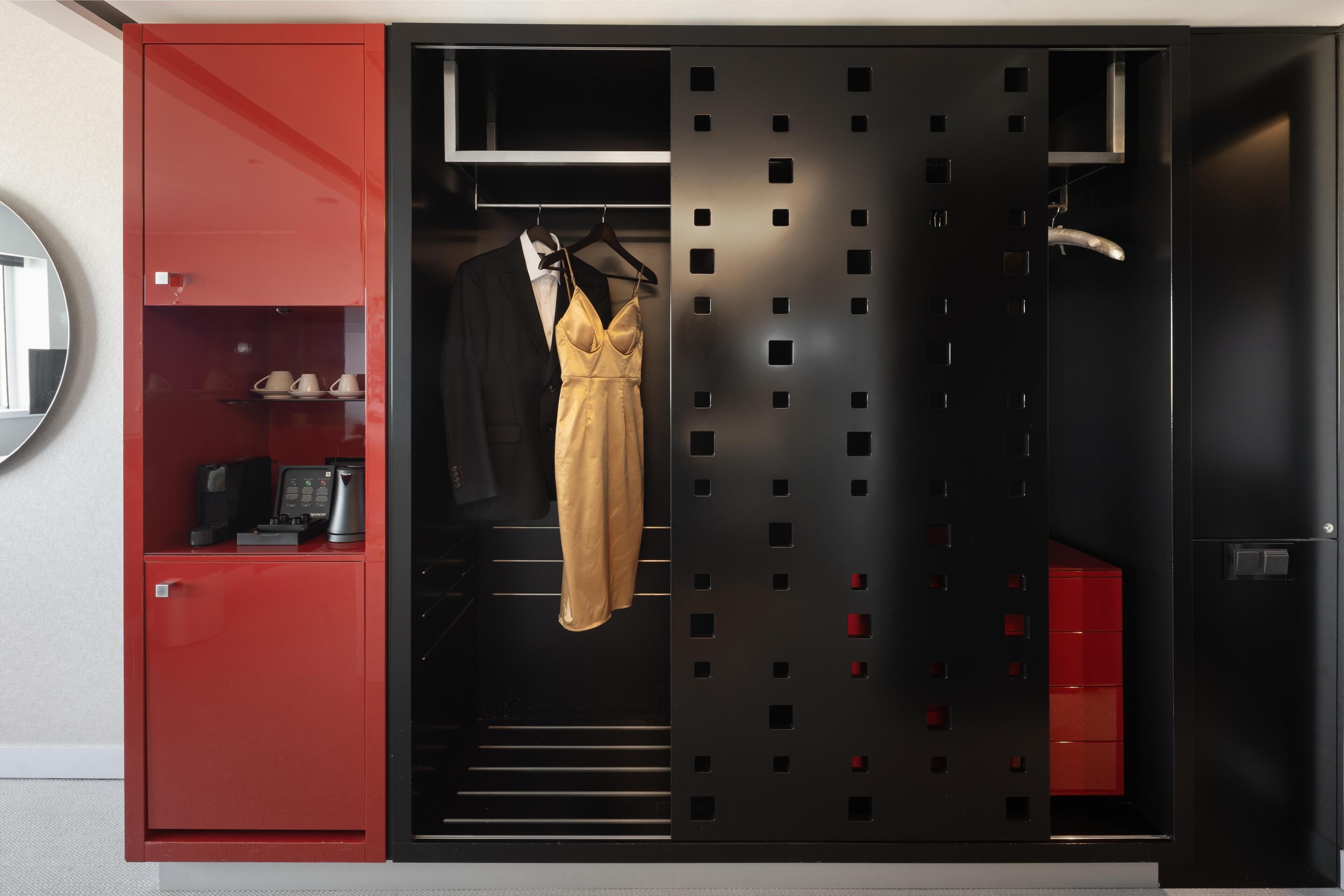 Habitación de hotel con un armario rojo, un mini bar negro y rojo, y un separador negro con espejo.