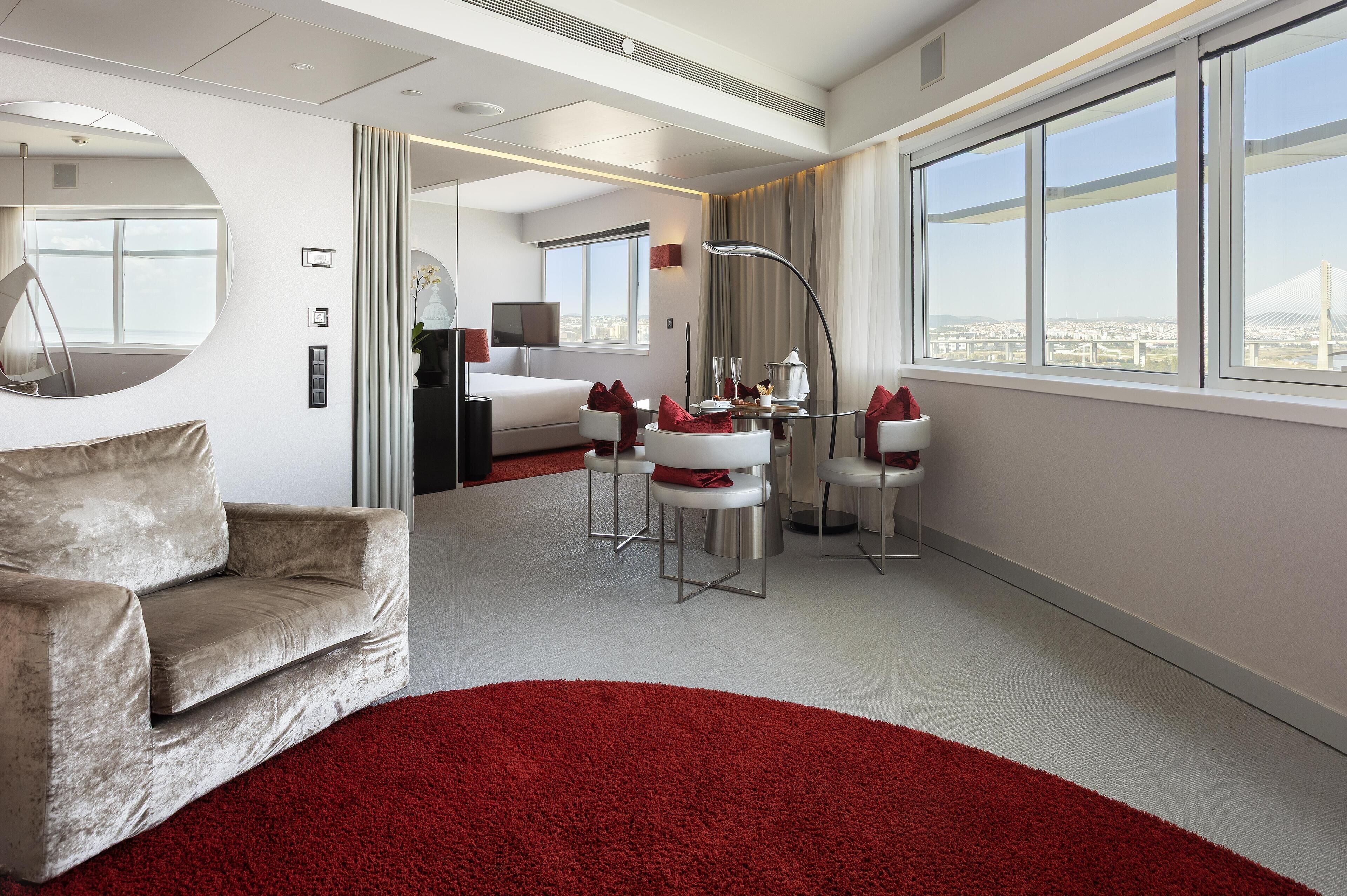 Quarto de hotel moderno com uma cama grande, um espaço de estar com tapete vermelho, um espelho redondo e vista para uma ponte.