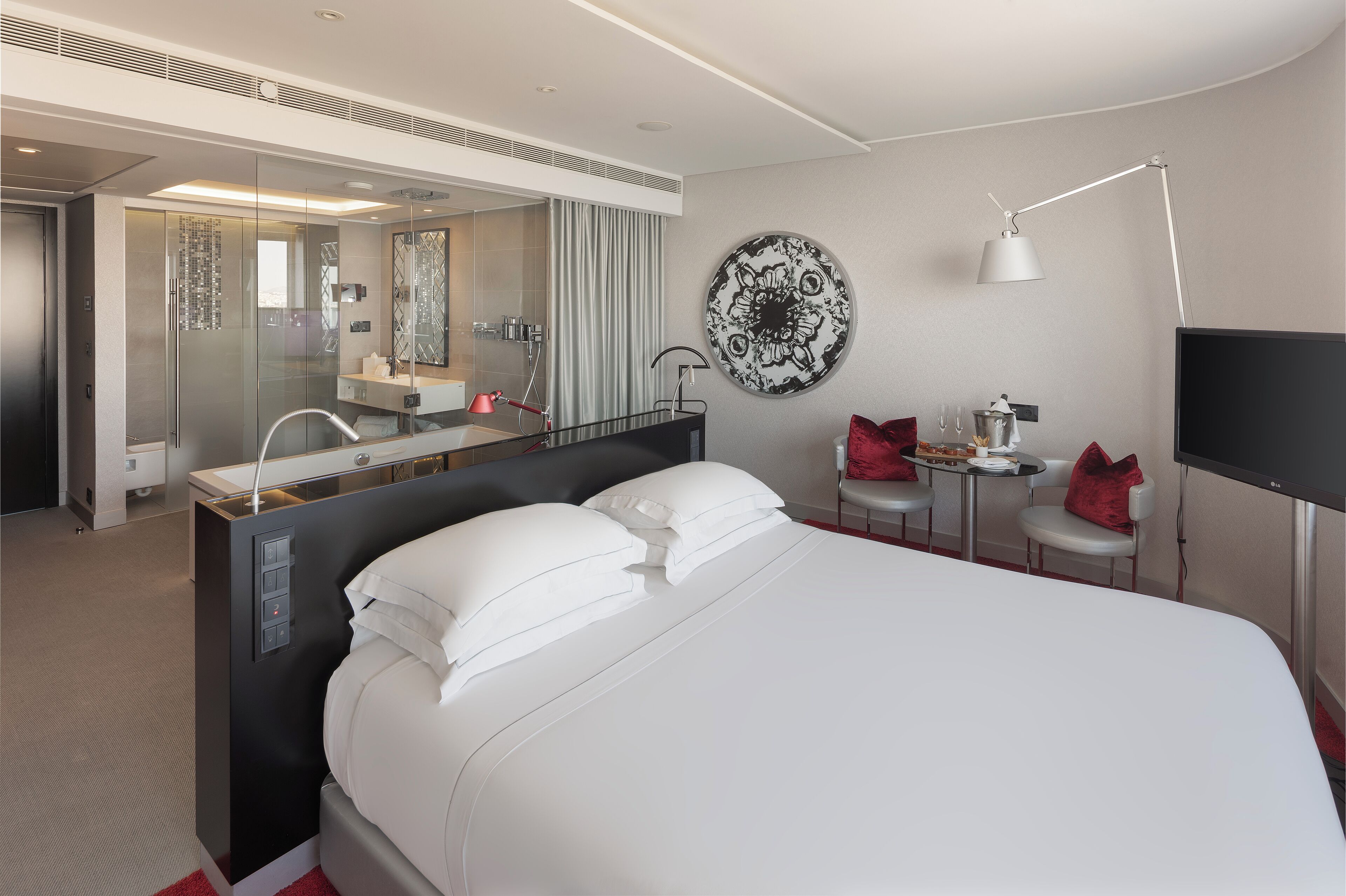 Una habitación de hotel moderna con una cama grande, una ducha con cerramiento de vidrio, un escritorio pequeño con una silla, un adorno redondo en la pared y un televisor de pantalla plana.