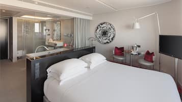 Una habitación de hotel moderna con una cama grande, una ducha con cerramiento de vidrio, un escritorio pequeño con una silla, un adorno redondo en la pared y un televisor de pantalla plana.