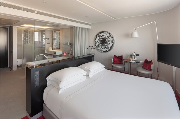 A modern hotel room with a large bed, a glass-enclosed shower, a small desk with a chair, a round wall decoration, and a flat-screen TV.