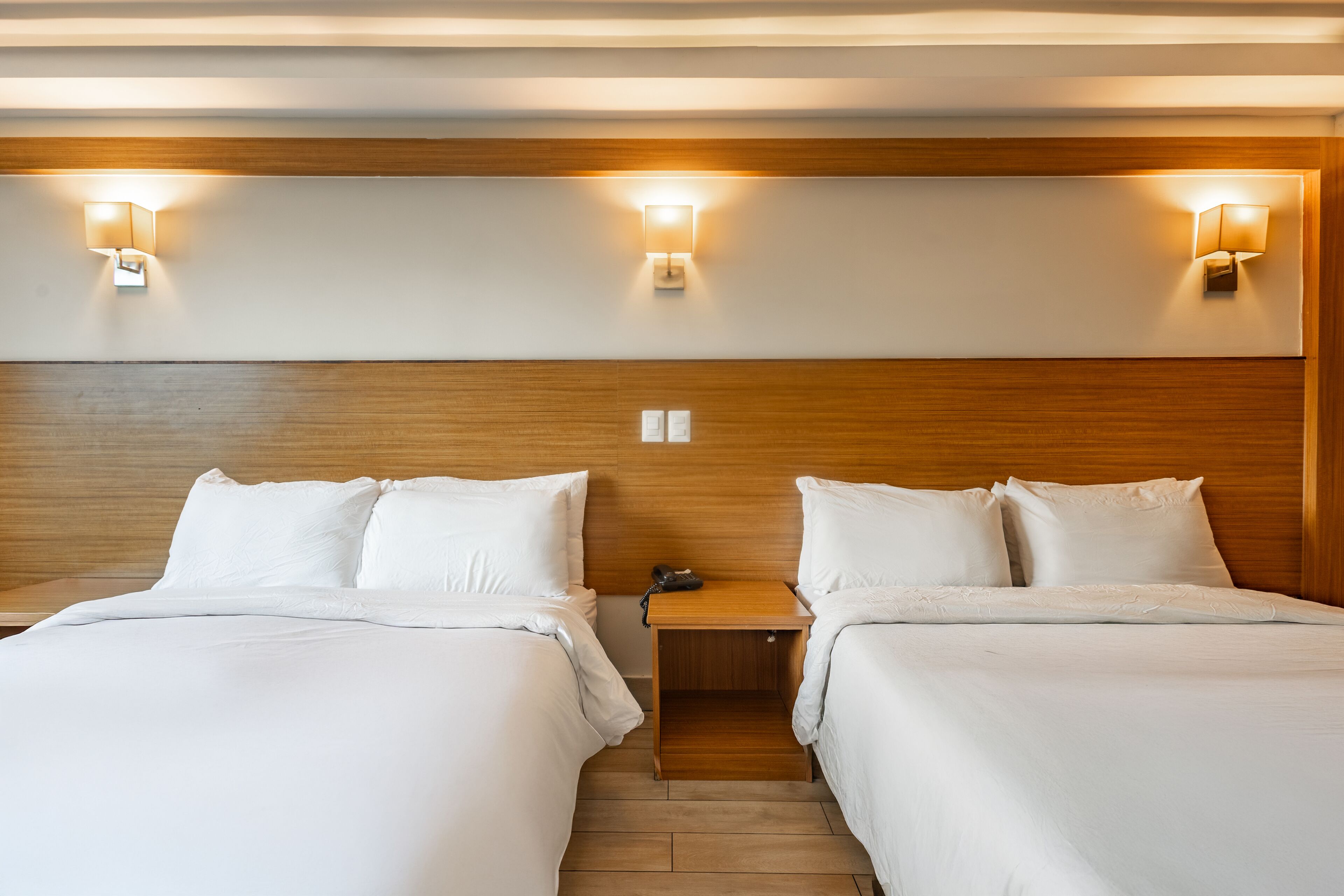 A hotel room with two beds, wooden headboard, bedside lamps, and a nightstand with a phone.