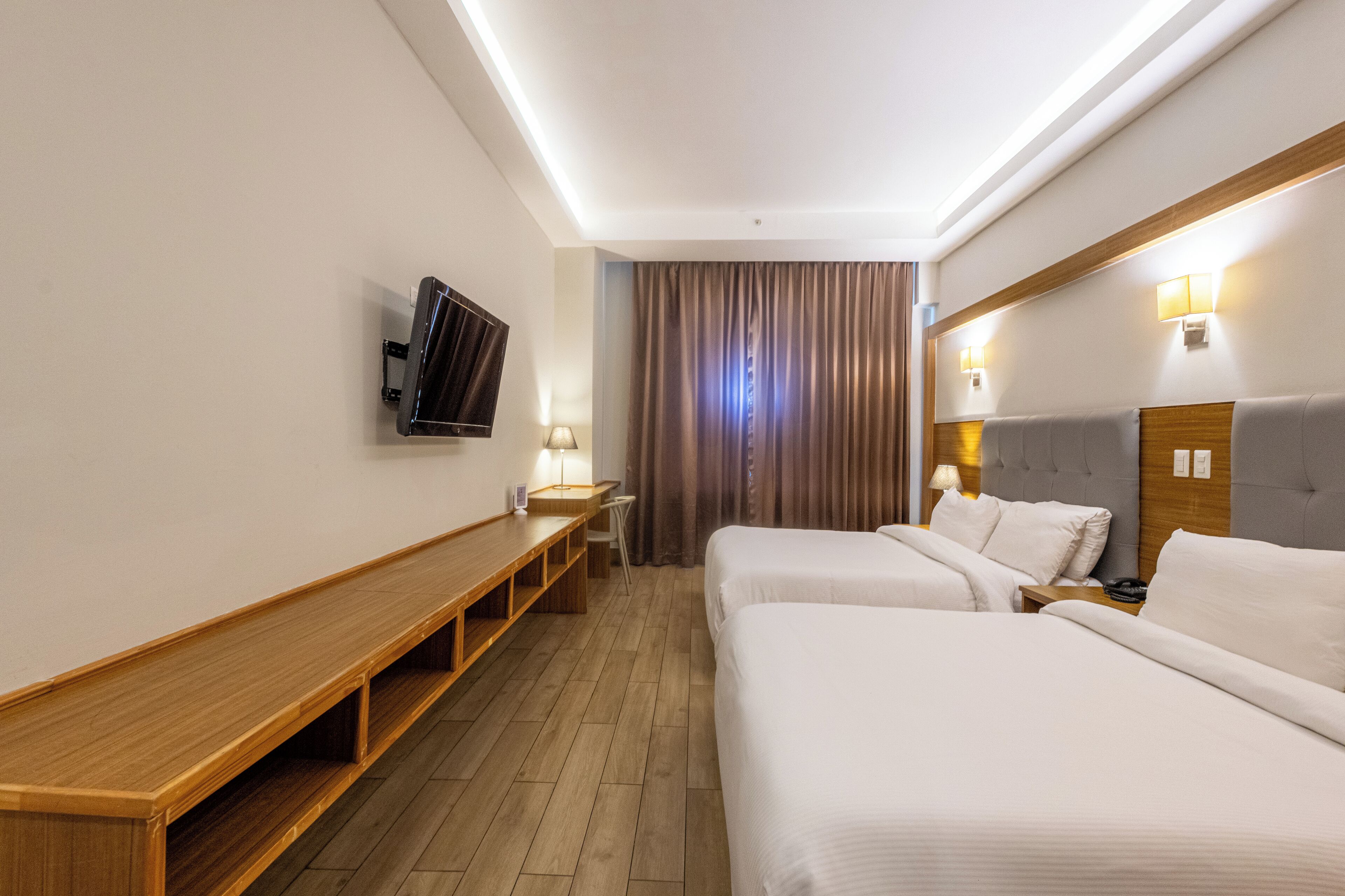 A hotel room with two beds, a wooden bench, a TV, and a desk with a lamp.