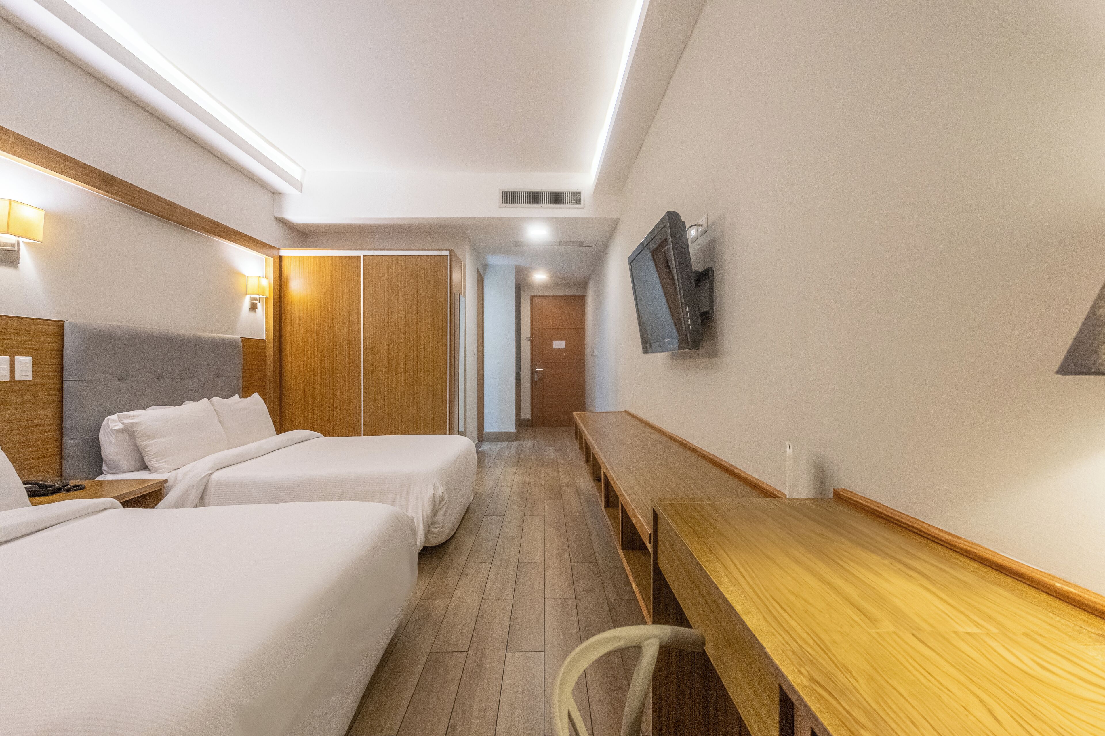 Habitación de hotel con dos camas, un escritorio de madera, un televisor montado en la pared y un armario de madera.