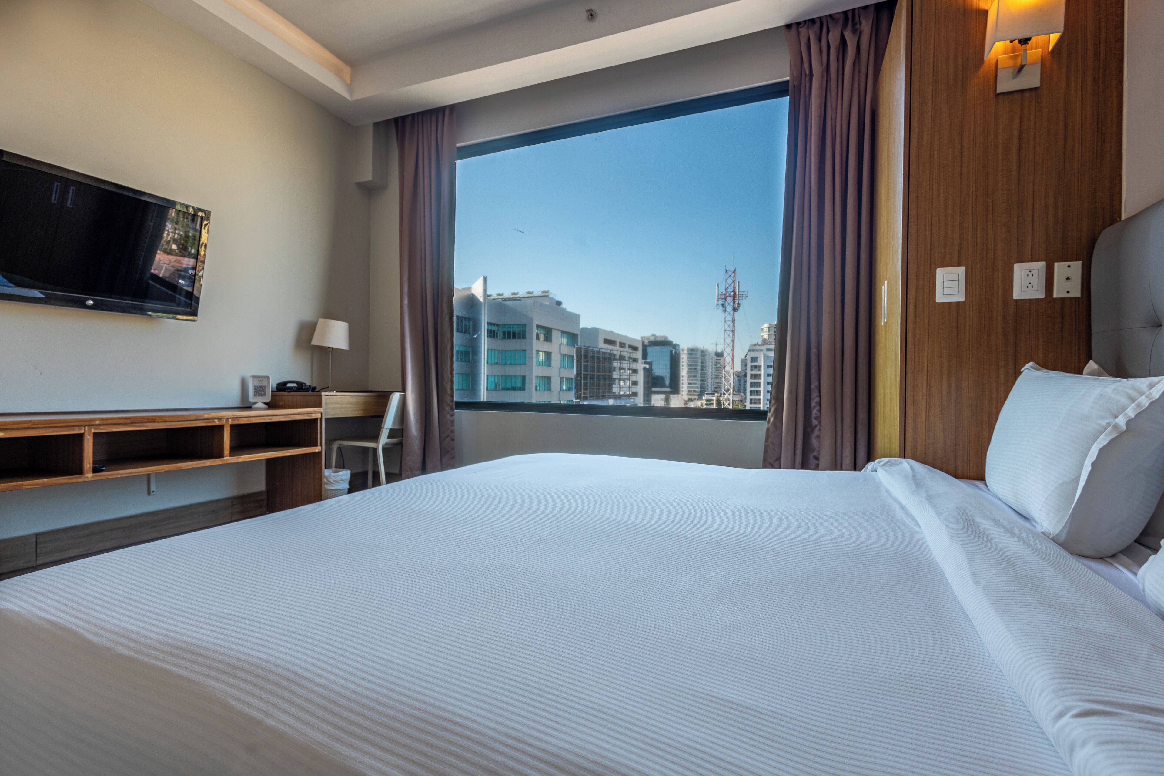 Quarto de hotel com uma cama grande, uma escrivaninha, uma televisão e uma janela com vista para a cidade.