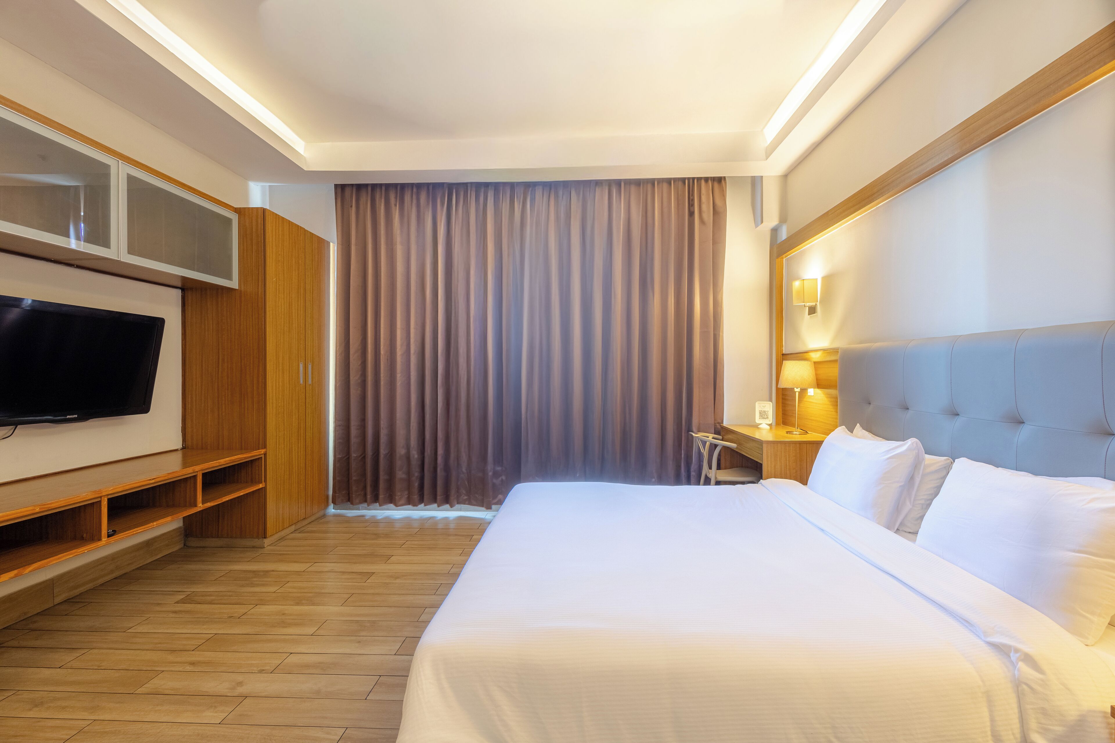 Habitación de hotel con una cama grande, un televisor montado en la pared, un escritorio con lámpara y una ventana cubierta por cortinas.