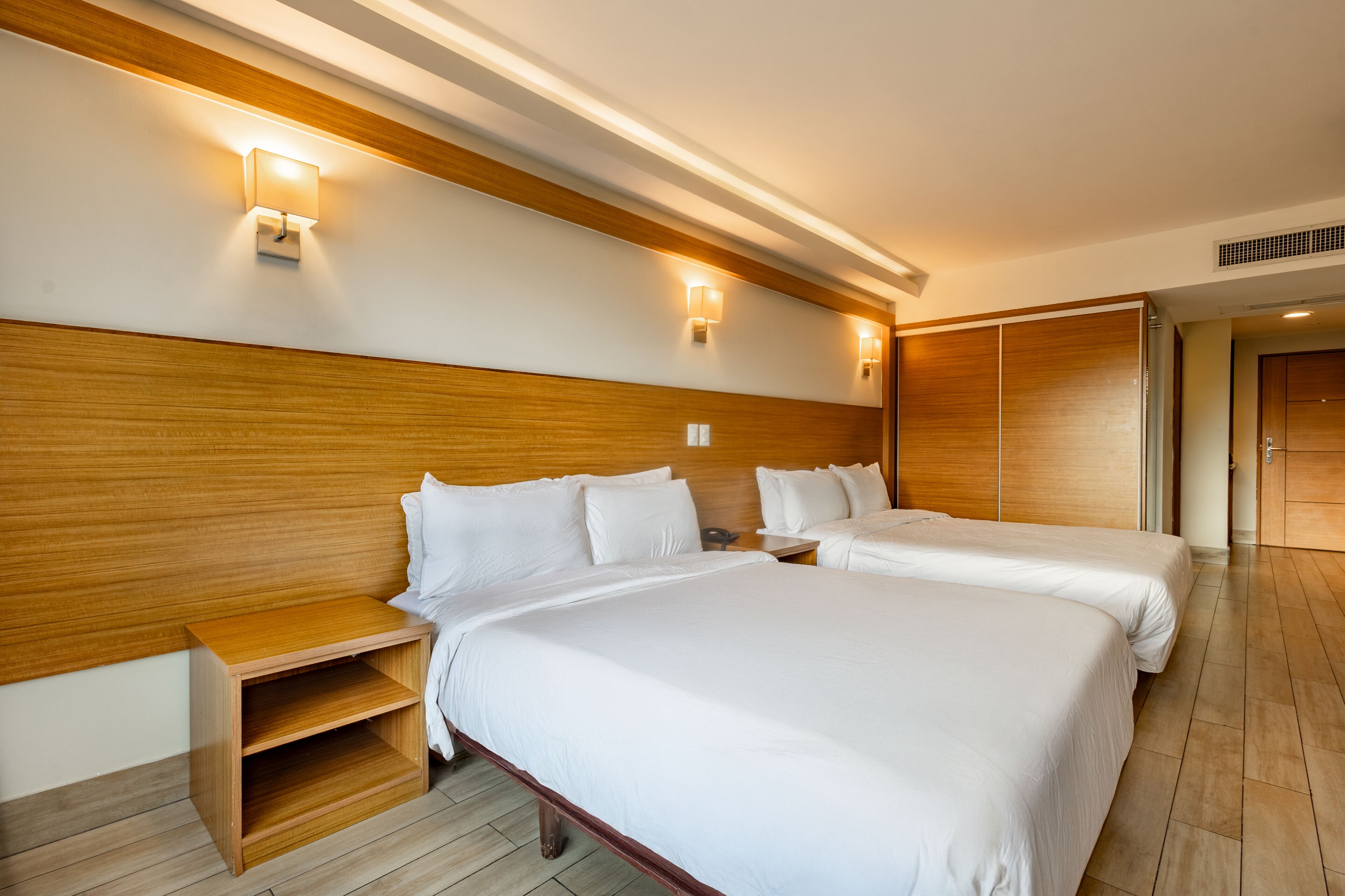 Una habitación de hotel con dos camas, cabecero de madera, mesitas de noche e iluminación empotrada.