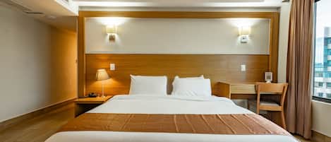 Quarto de hotel com uma cama grande, uma mesa de trabalho, duas luminárias na parede e vista da cidade pela janela.