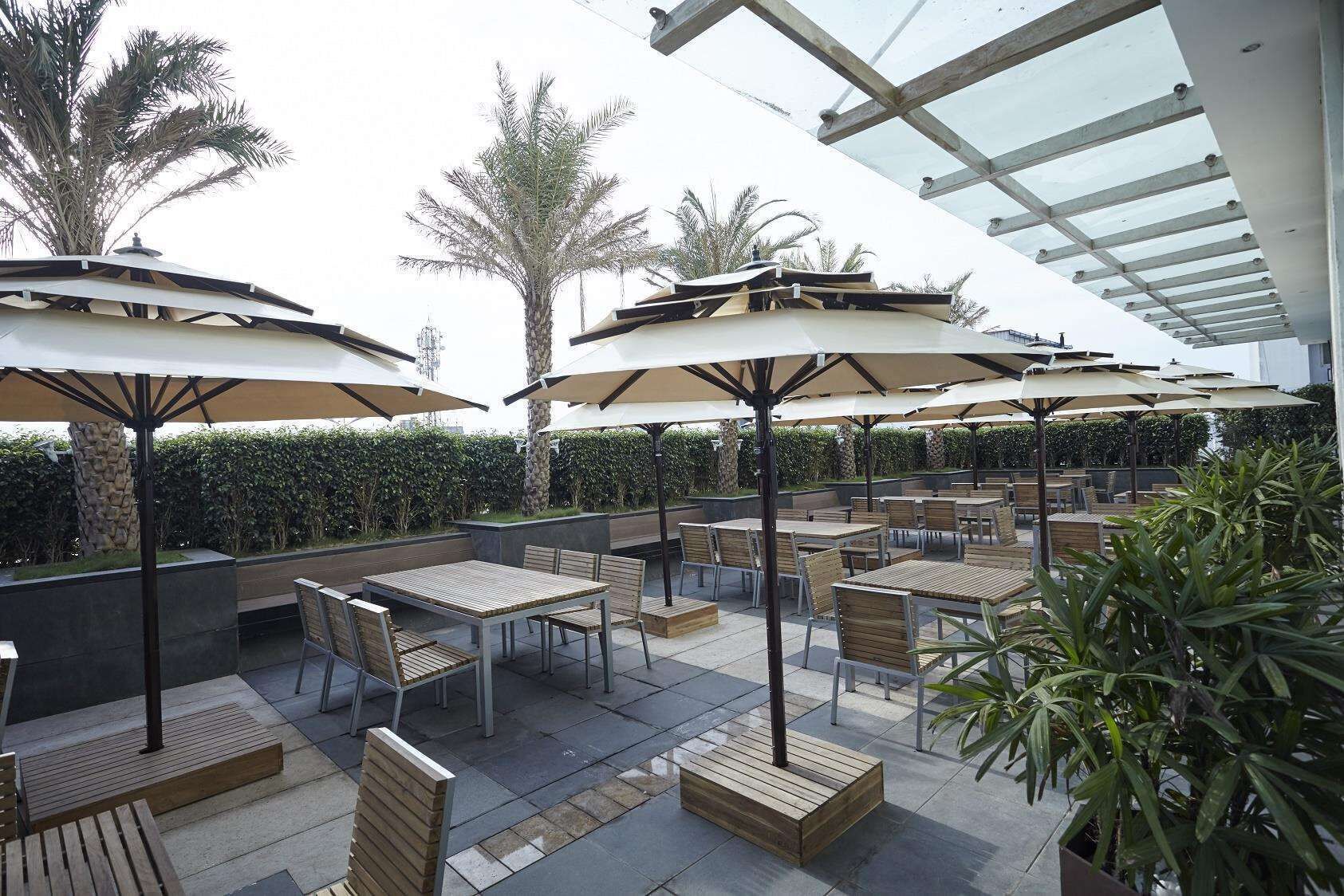 An outdoor patio area with multiple tables and chairs, shaded by umbrellas, and surrounded by palm trees and greenery.