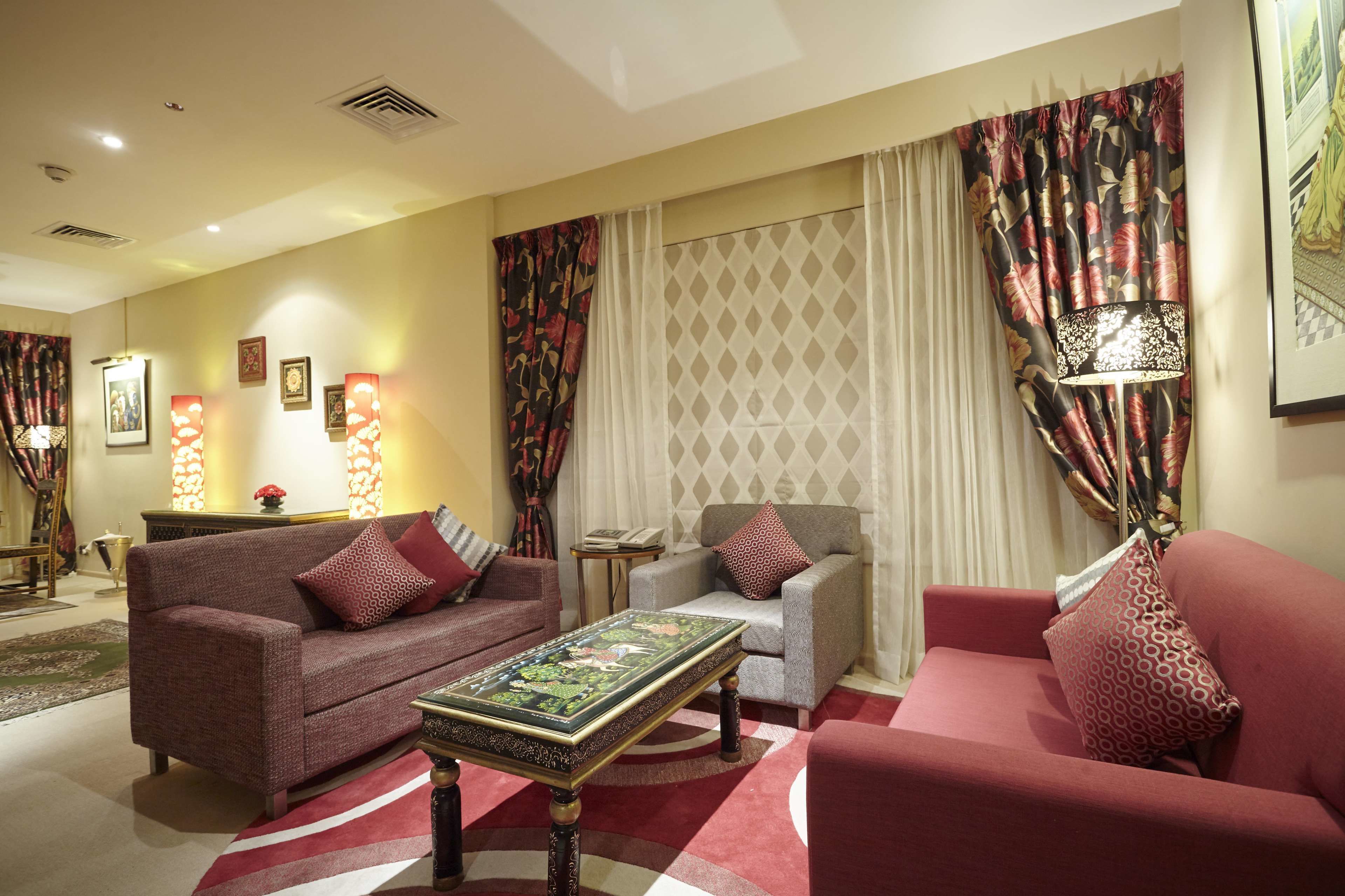 Un salon avec un canapé, un fauteuil et une table basse. On y trouve des rideaux, une lampe et des tableaux encadrés accrochés au mur.