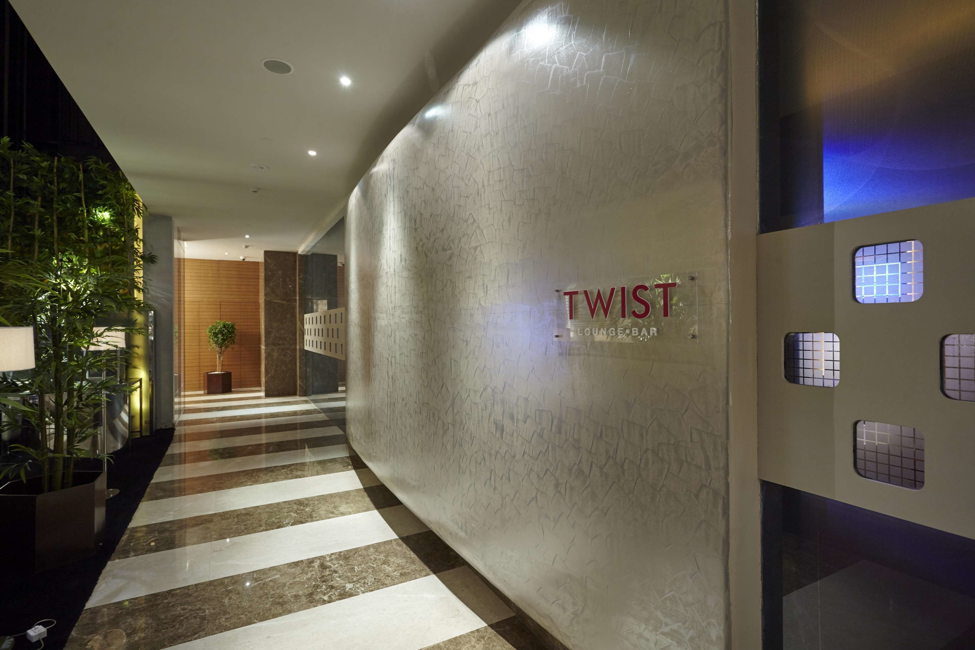 Un vestíbulo moderno con piso de mármol pulido, una pared grande con el nombre 'TWIST' y 'Lounge Bar' a la vista, y una planta en maceta al frente.