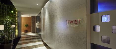 A modern lobby with a polished marble floor, a large wall with the name 'TWIST' and 'Lounge Bar' visible, and a potted plant in the foreground.