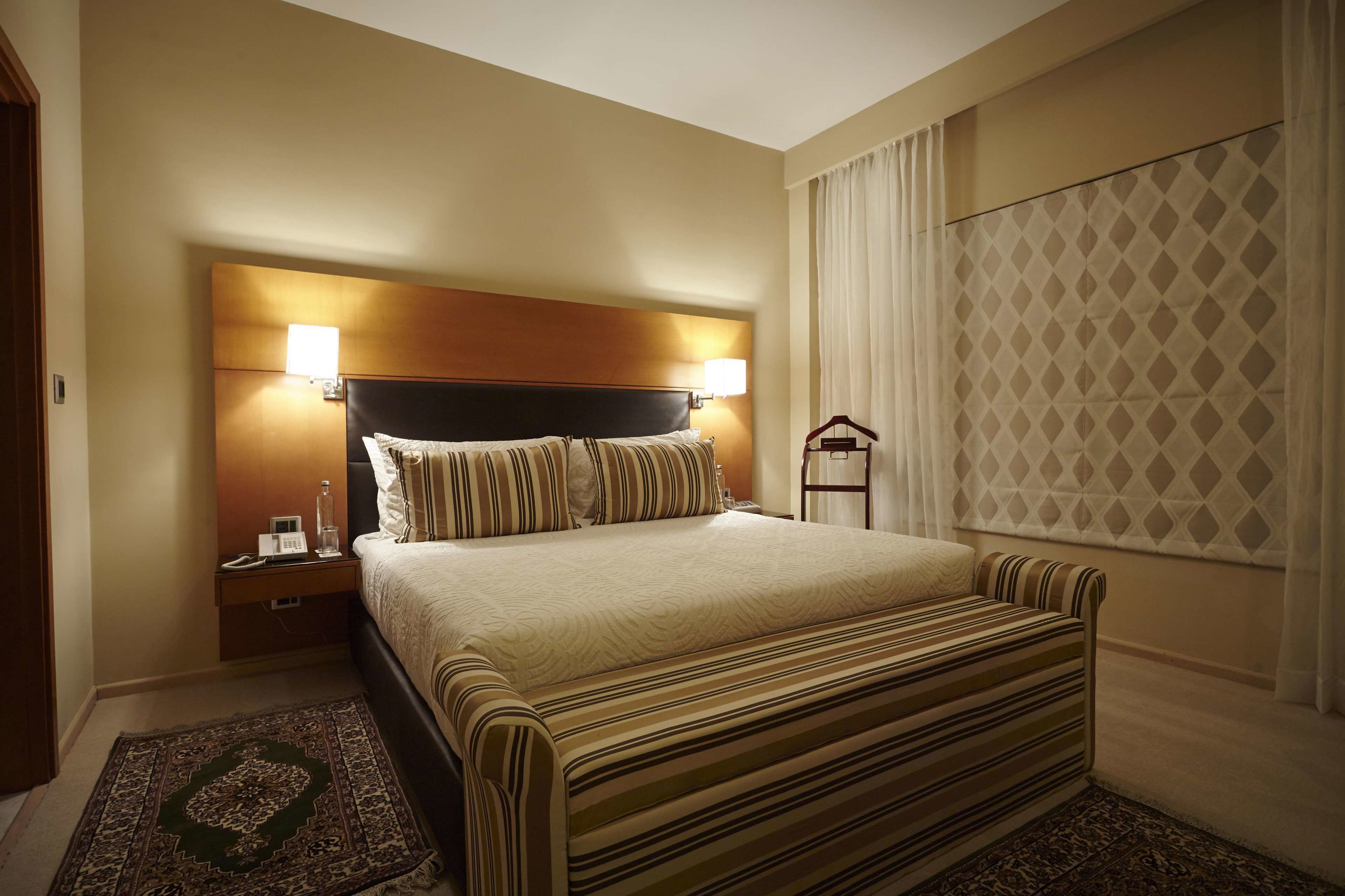 A hotel room with a large bed, two bedside lamps, a nightstand, a window with curtains, and a patterned rug.