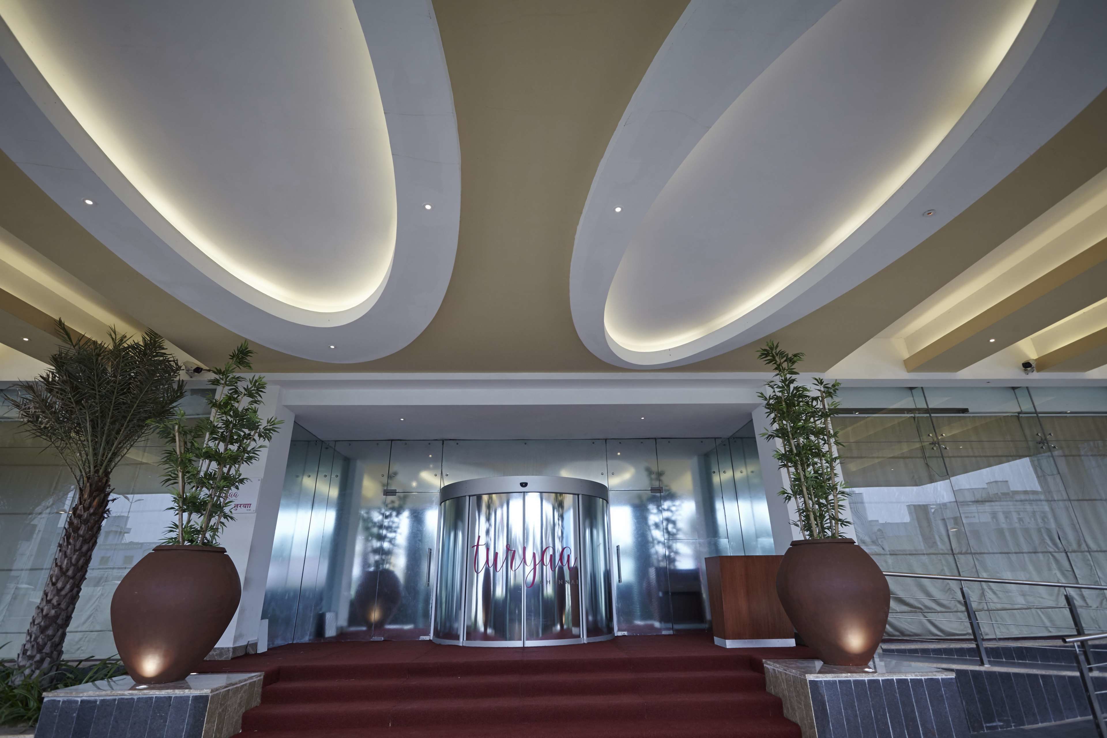 A modern hotel lobby with a revolving door, red carpeted stairs, and large potted plants.