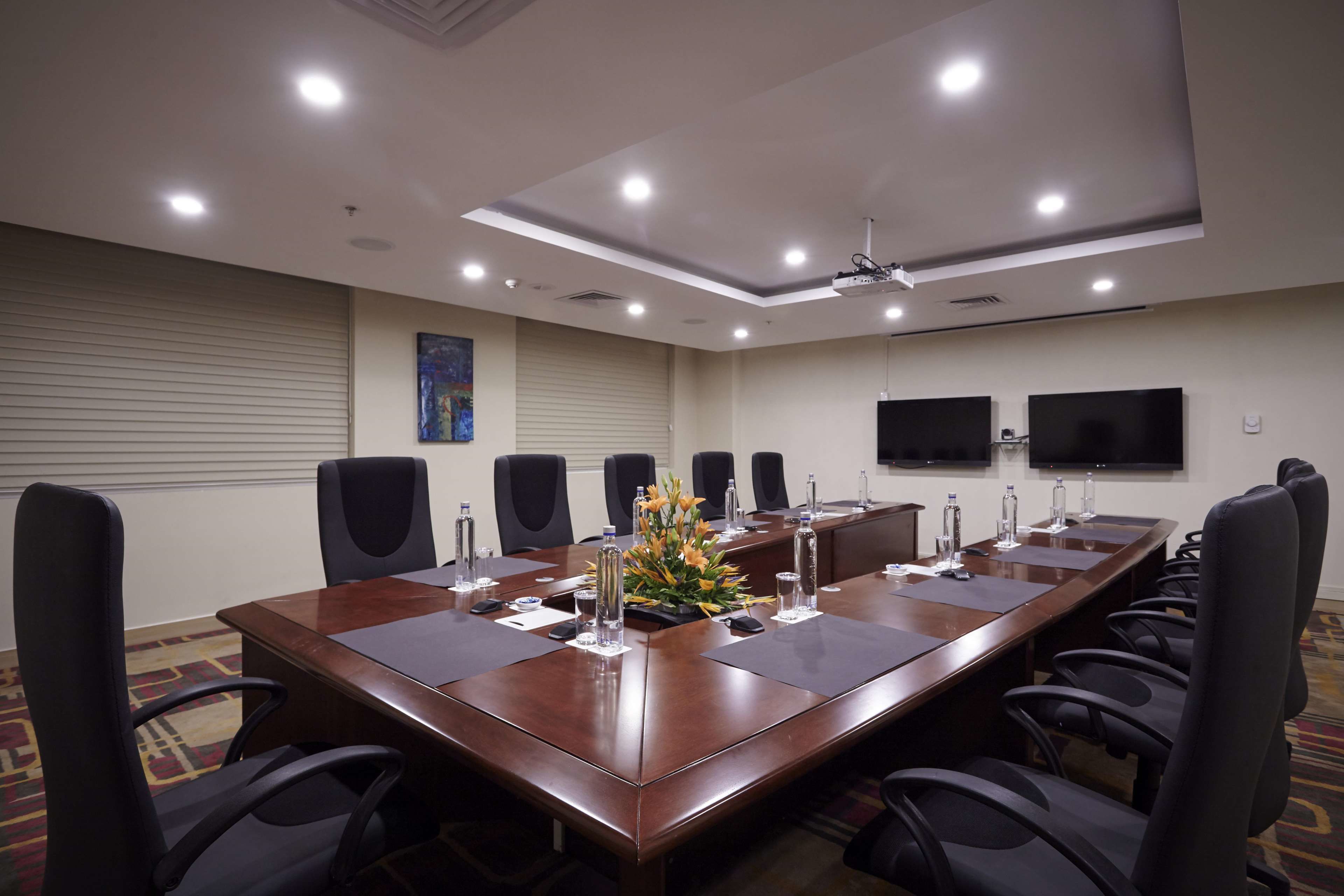 Un salón de conferencias con una larga mesa, sillas negras, botellas de agua y un centro de mesa con flores.
