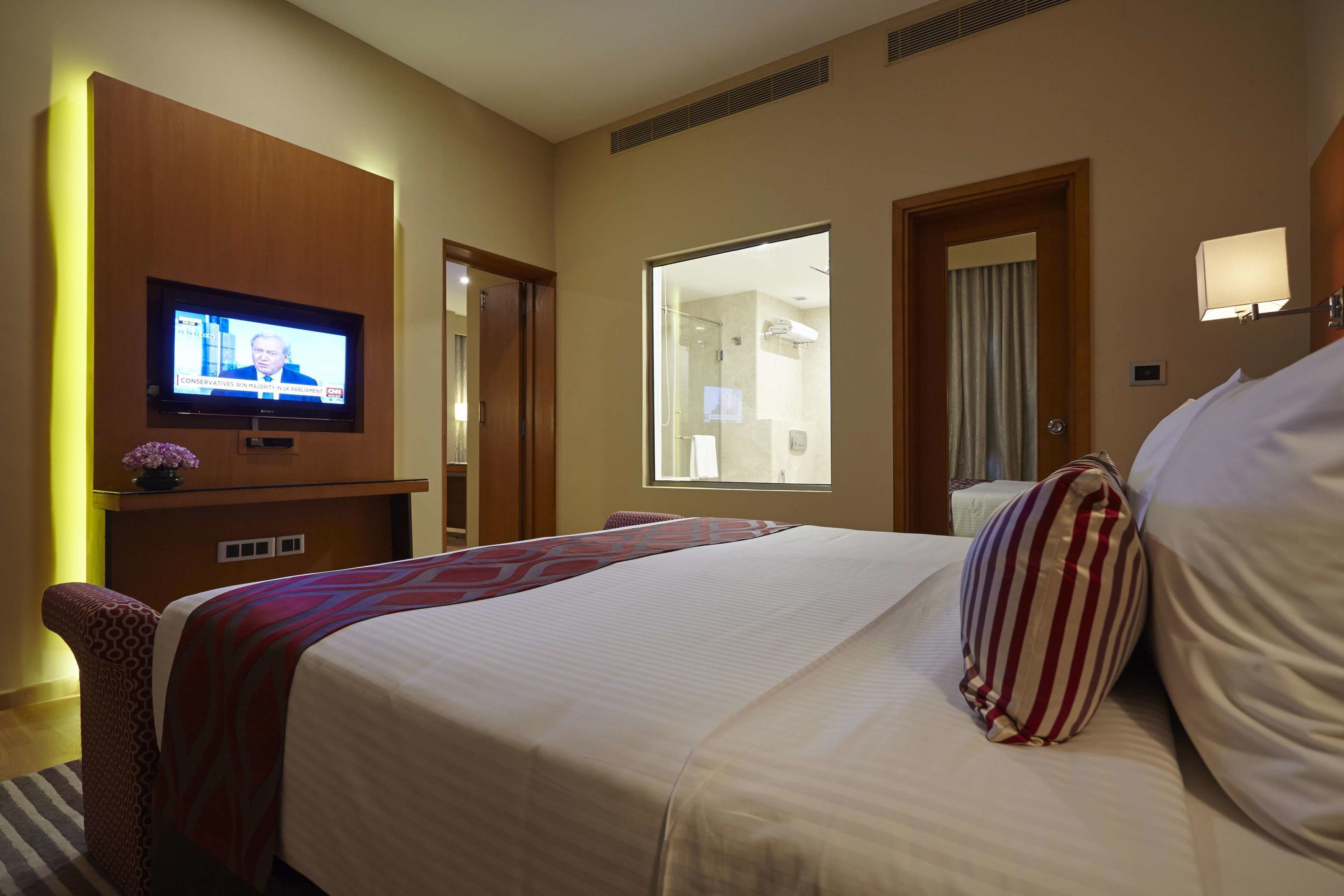 A hotel room with a large bed, a television mounted on a wooden panel, a window, and a bathroom visible through the door.