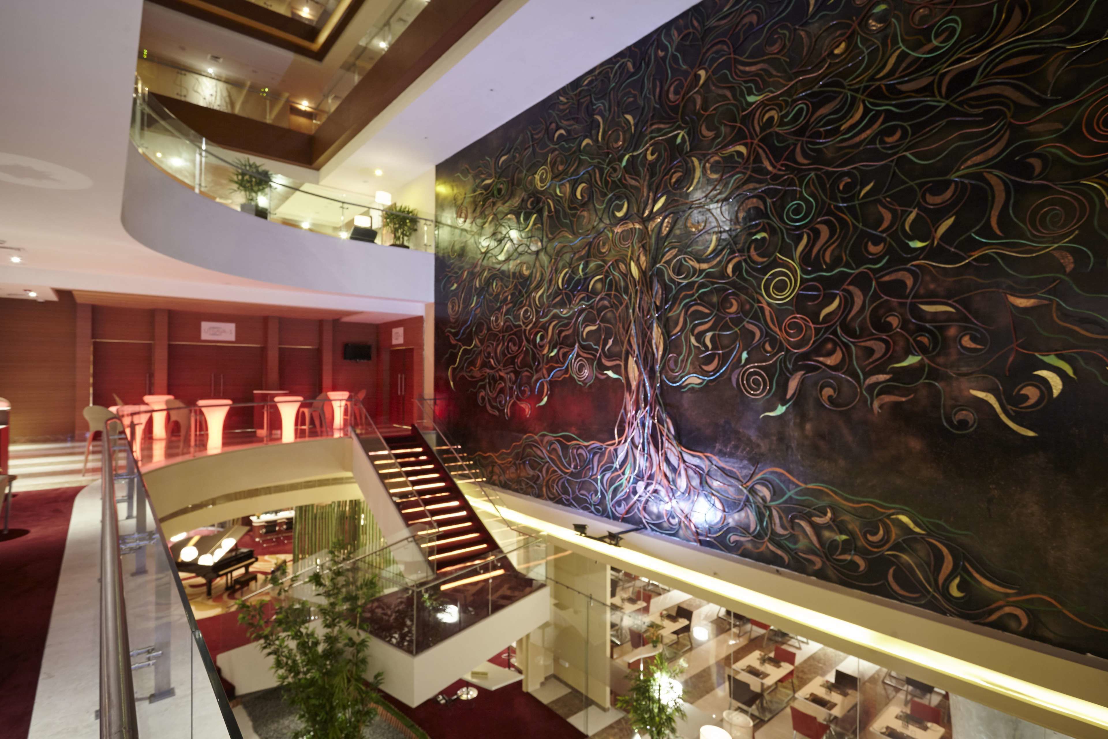 A multi-level hotel interior with a large, abstract mural on the upper wall, a staircase connecting floors, and a seating area with tables and chairs.