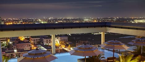 A rooftop pool area with umbrellas and lounge chairs, overlooking a cityscape at night.