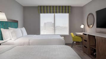Ett hotellrum med två sängar, ett skrivbord, en stol, en TV och ett fönster med utsikt över byggnader.
