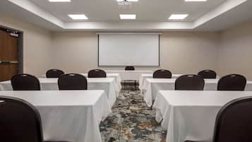 Una sala de conferencias con mesas blancas, sillas oscuras, piso alfombrado, una pantalla de proyector y una pizarra.