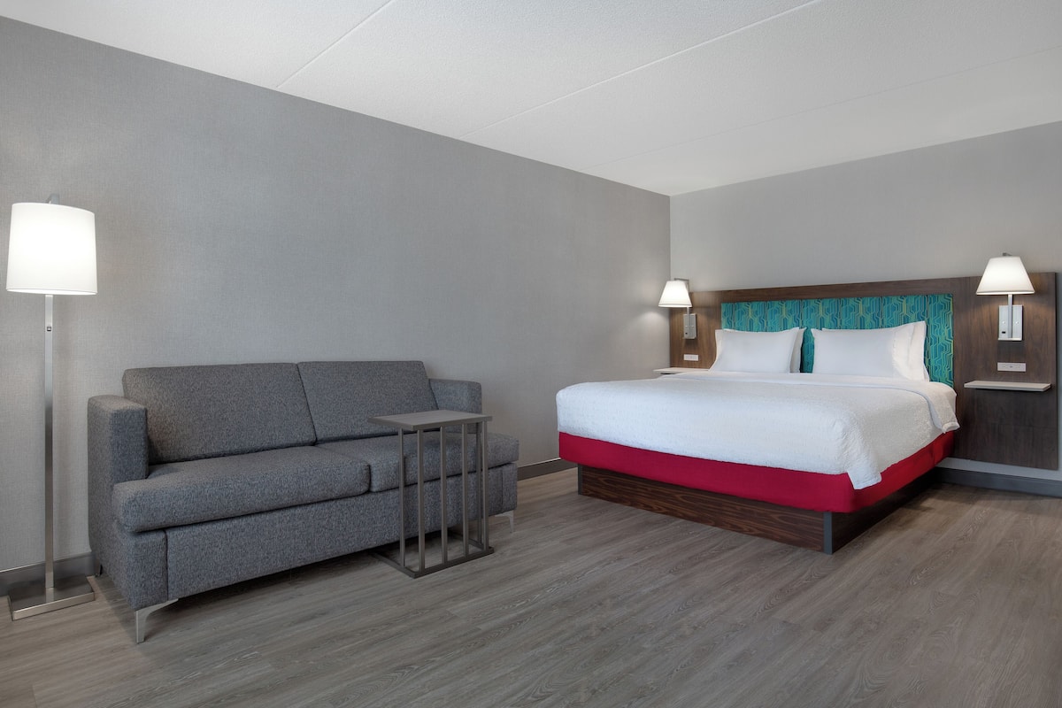 Hotellihuone, jossa on harmaa sohva, pieni pöytä, punaisella alustalla varustettu sänky ja kaksi yöpöytävalaisinta.