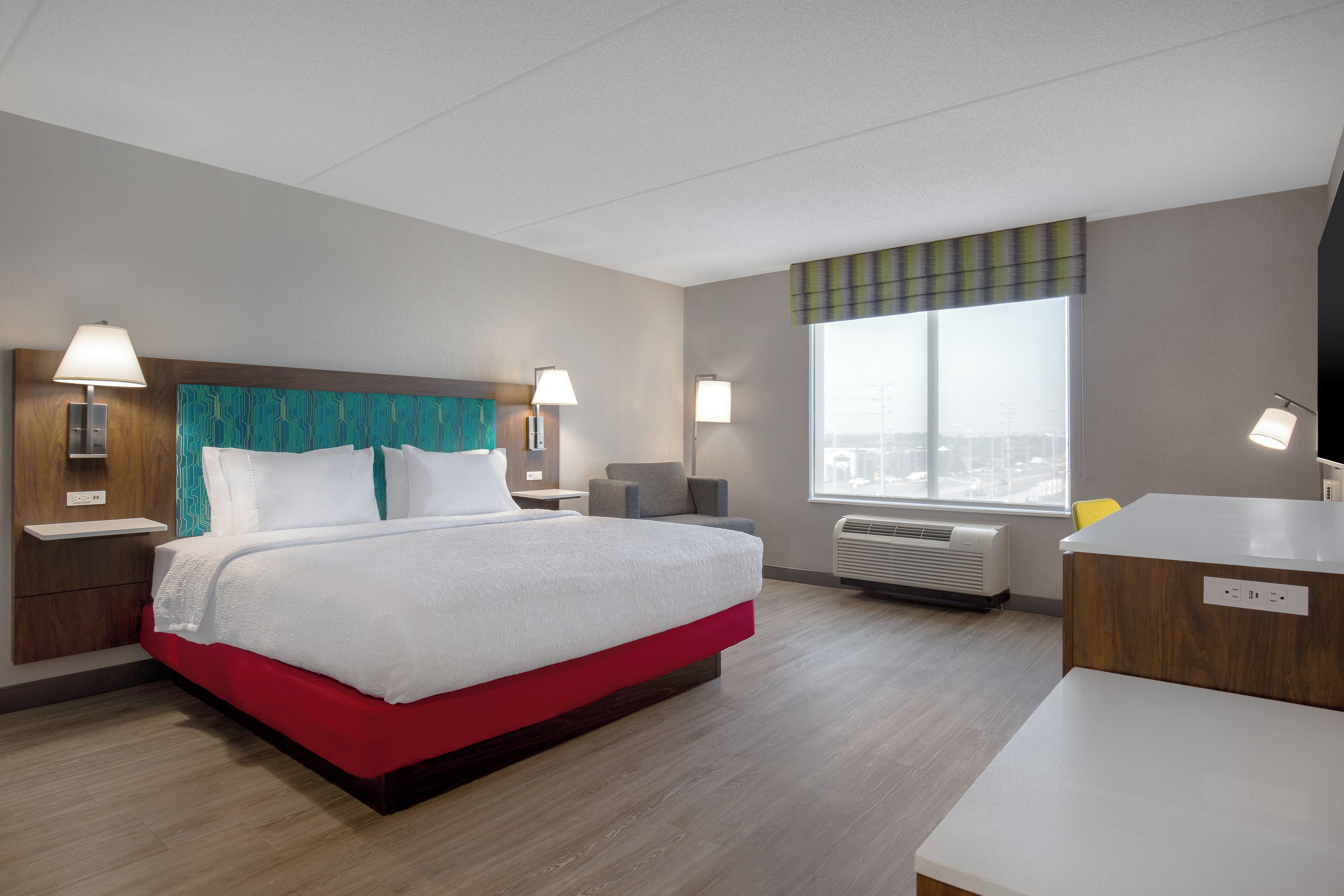 A hotel room with a bed, bedside lamps, a desk, a chair, and a large window with a view.