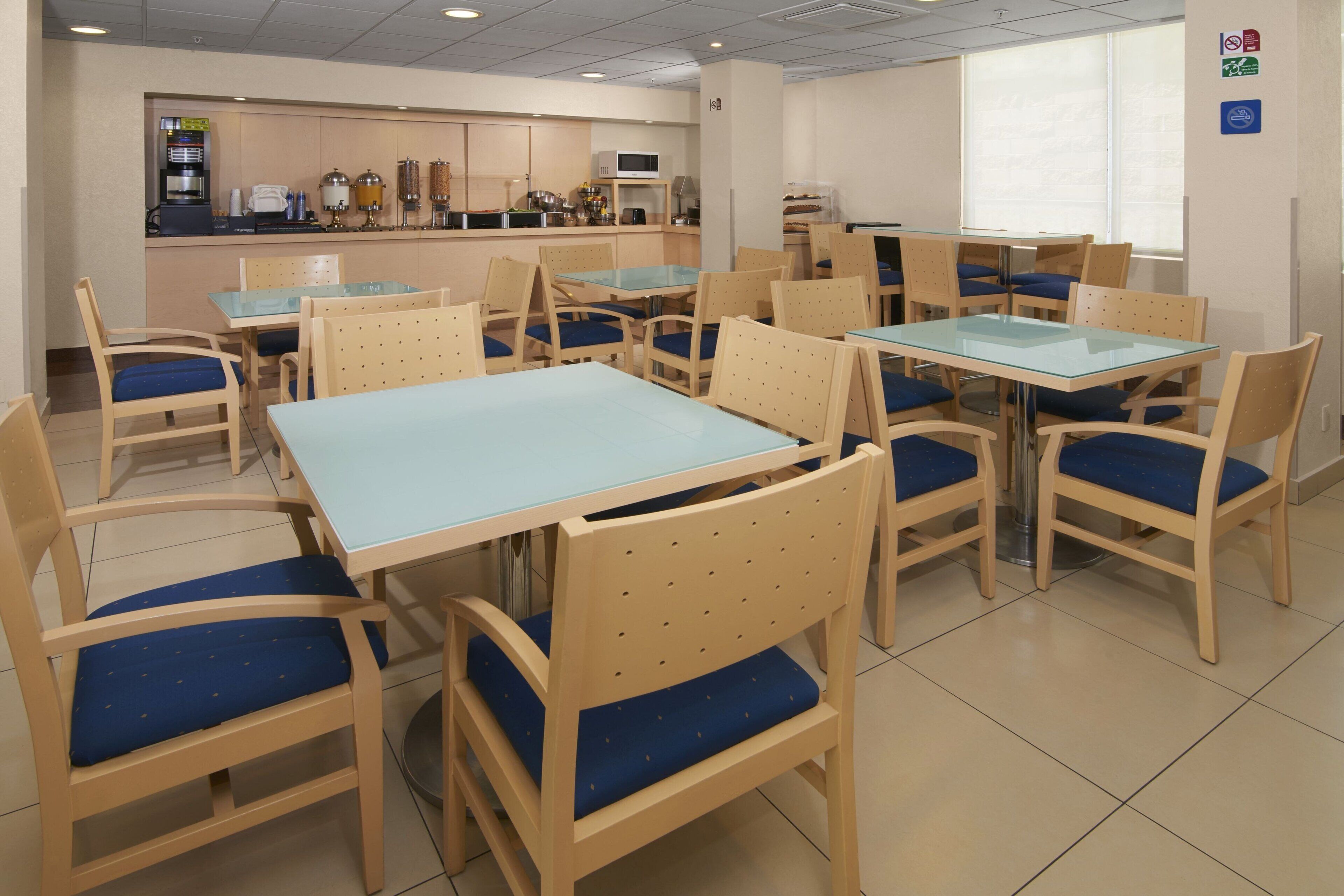 A dining area with multiple tables and chairs, a kitchen in the background, and a window with blinds.