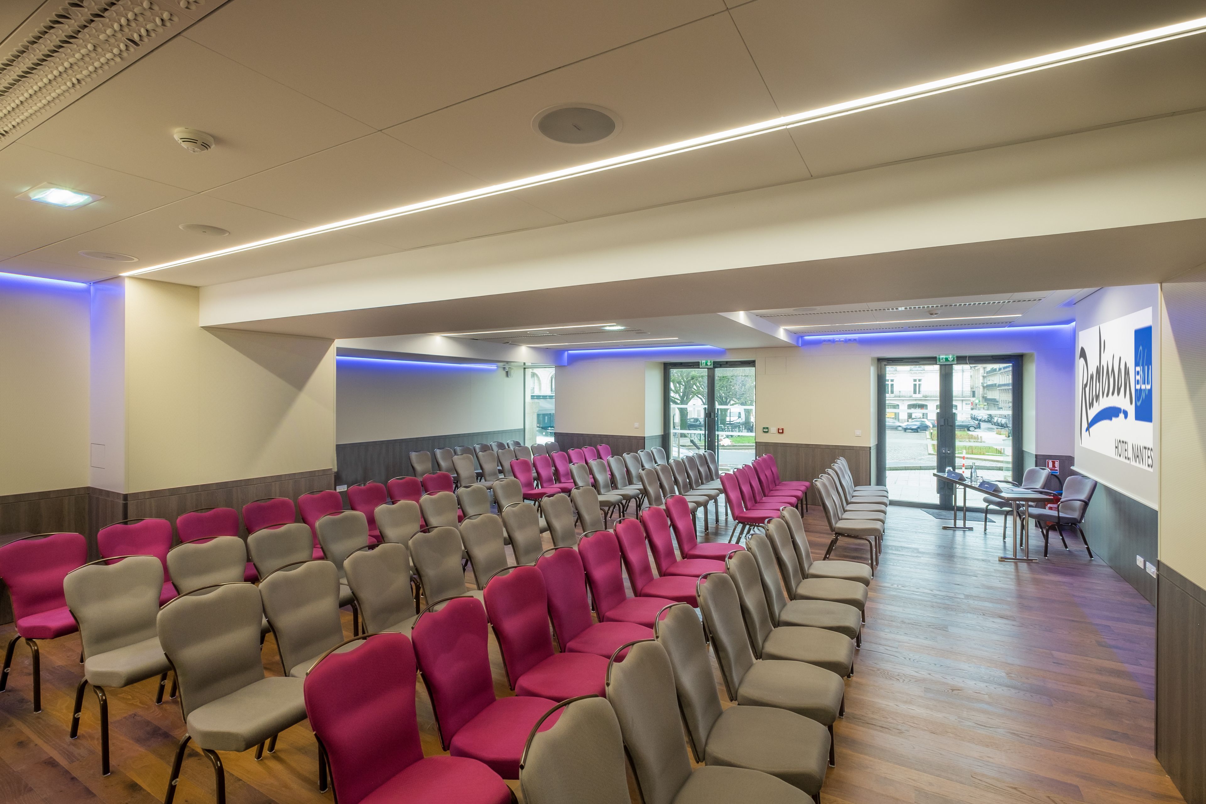 A conference room with rows of chairs, a wooden floor, and a view of the outside through large windows.