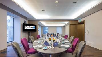 Un salón de conferencias con una mesa redonda, sillas y una pantalla de televisión mostrando el nombre del Hotel Radisson.