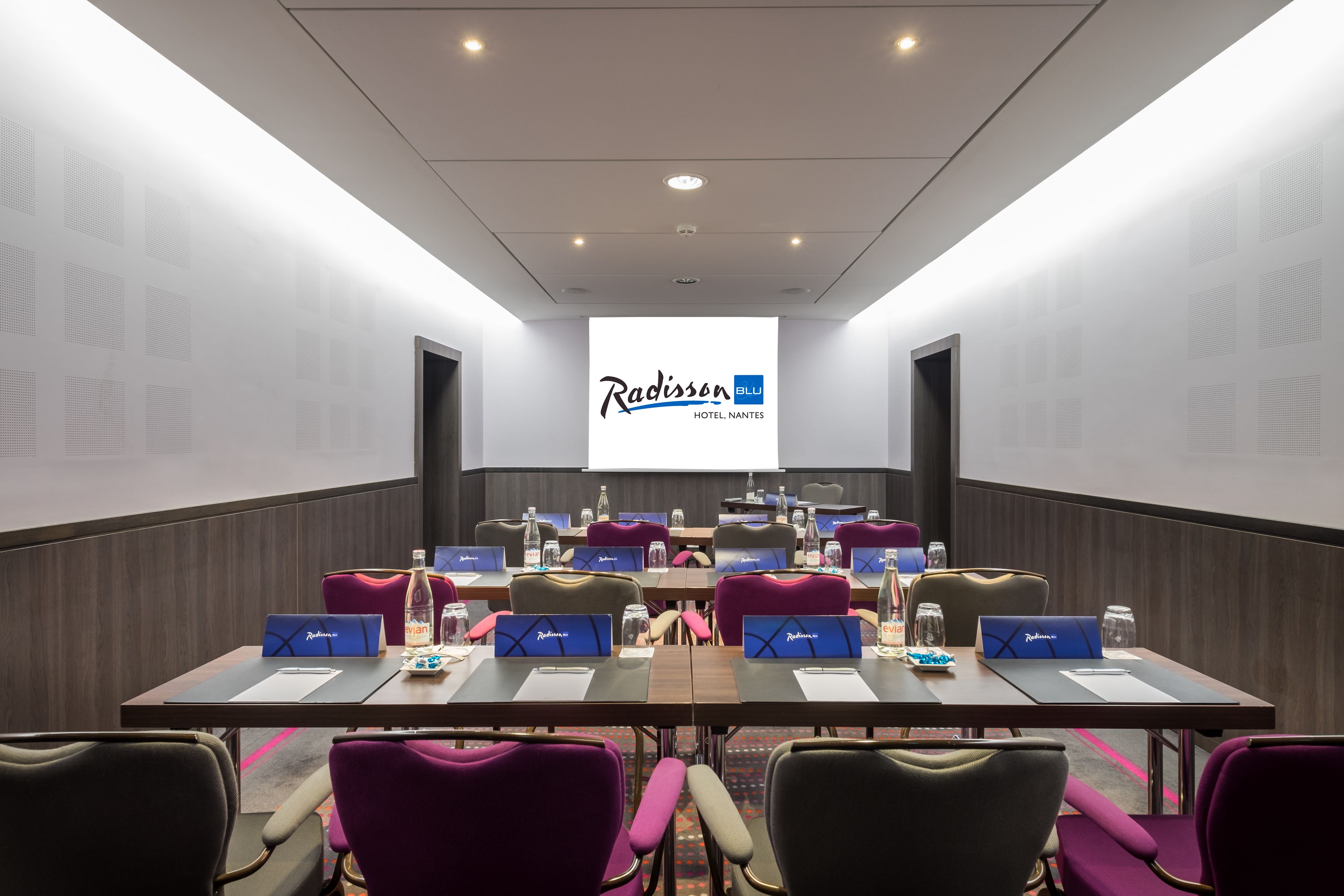 Une salle de conférence avec une longue table, de nombreuses chaises, des plaques nominatives, des bouteilles d’eau et un écran de présentation affichant le logo Radisson ELU.