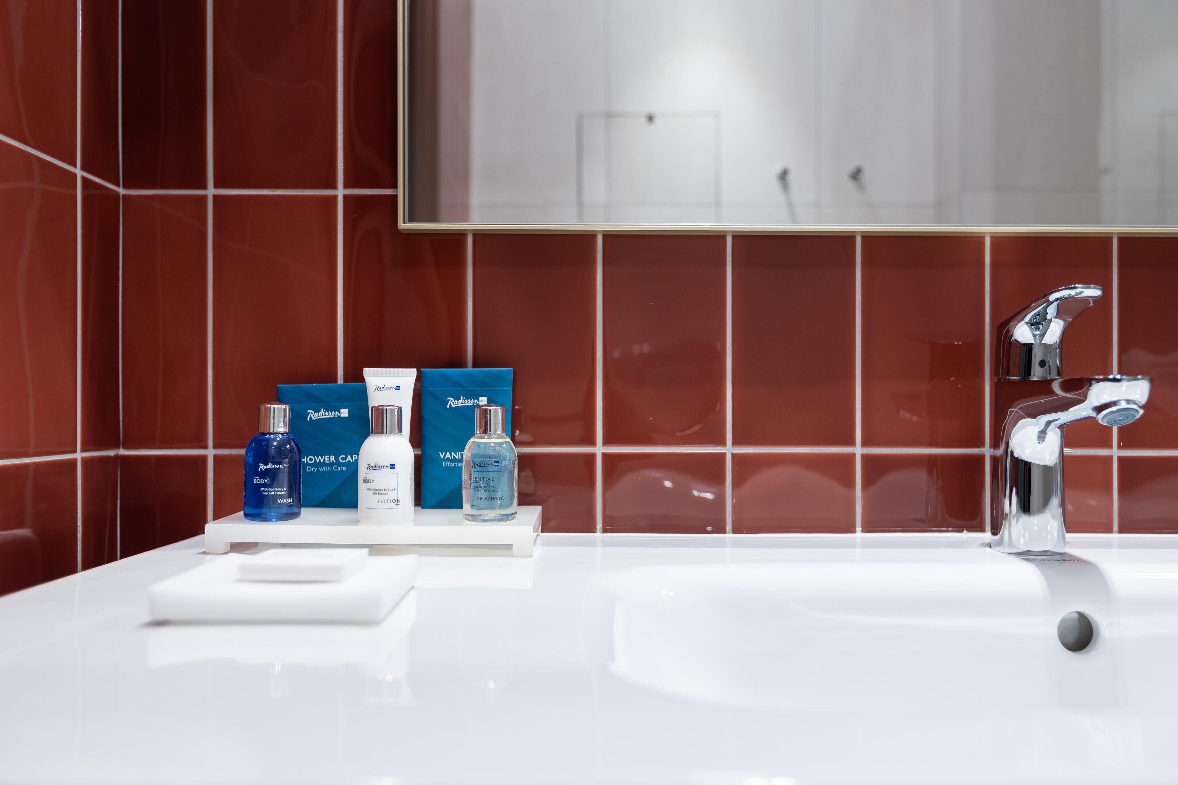 A bathroom with a white sink, a mirror, and red tiled walls. There are three bottles of shower gel and a bottle of shampoo on the sink.