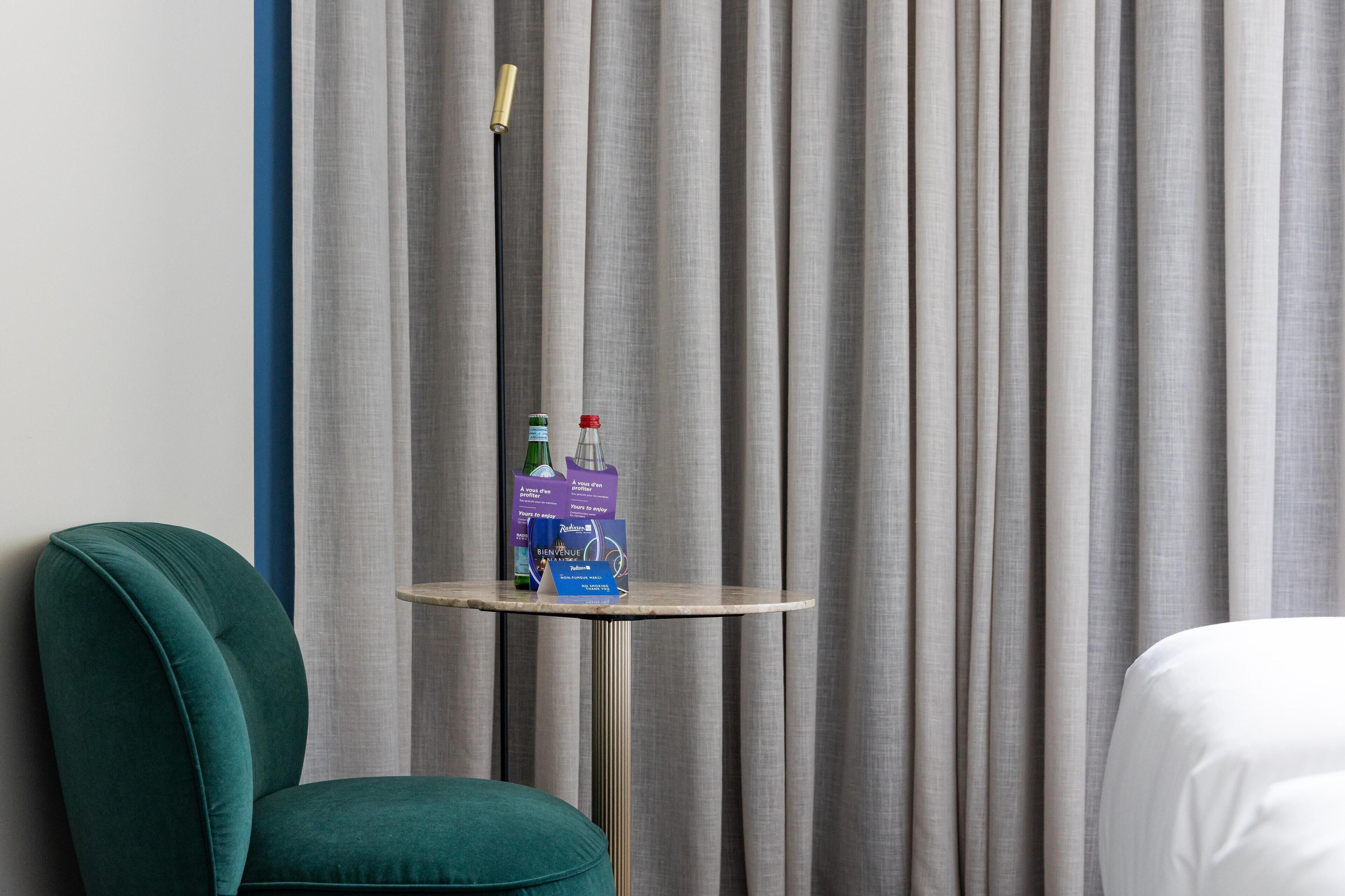 Una habitación de hotel con un sillón color turquesa, una mesita redonda con agua embotellada y un folleto, y una cama con ropa de cama blanca.