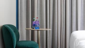 Una habitación de hotel con un sillón color turquesa, una mesita redonda con agua embotellada y un folleto, y una cama con ropa de cama blanca.