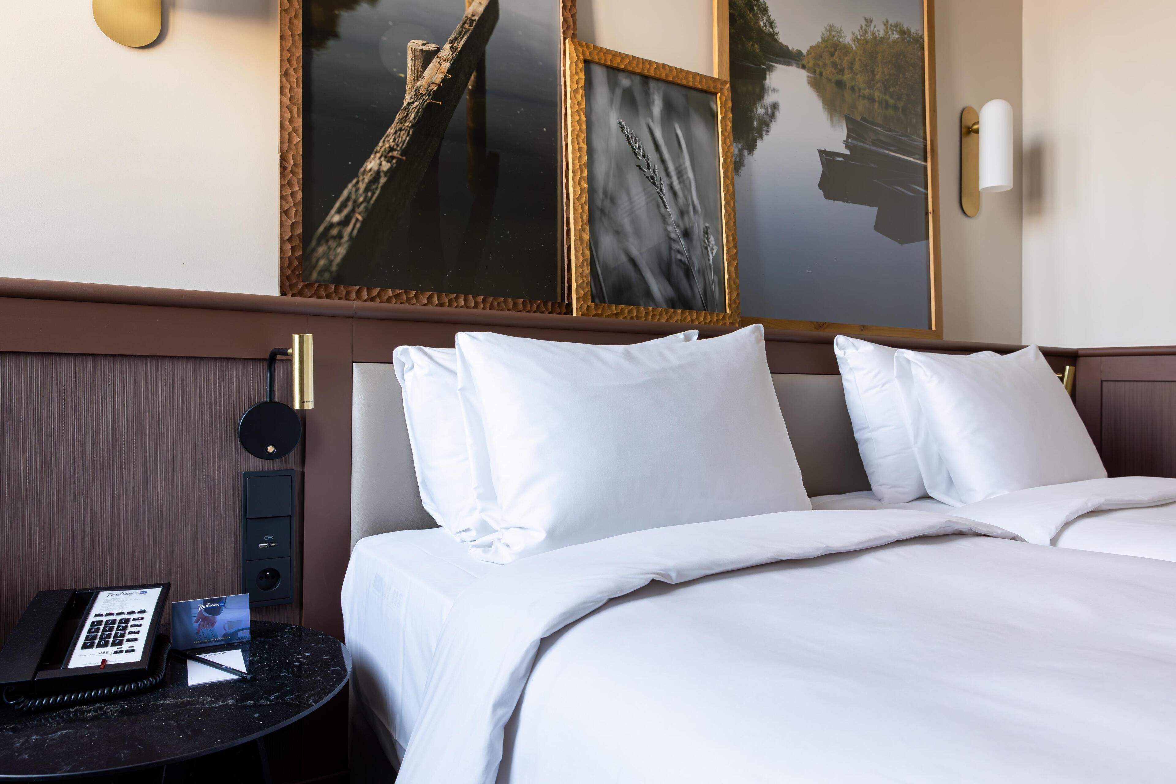Hotelkamer met een bed, witte kussens, een nachtkastje met een telefoon en een kaart, en een aan de muur hangend schilderij.